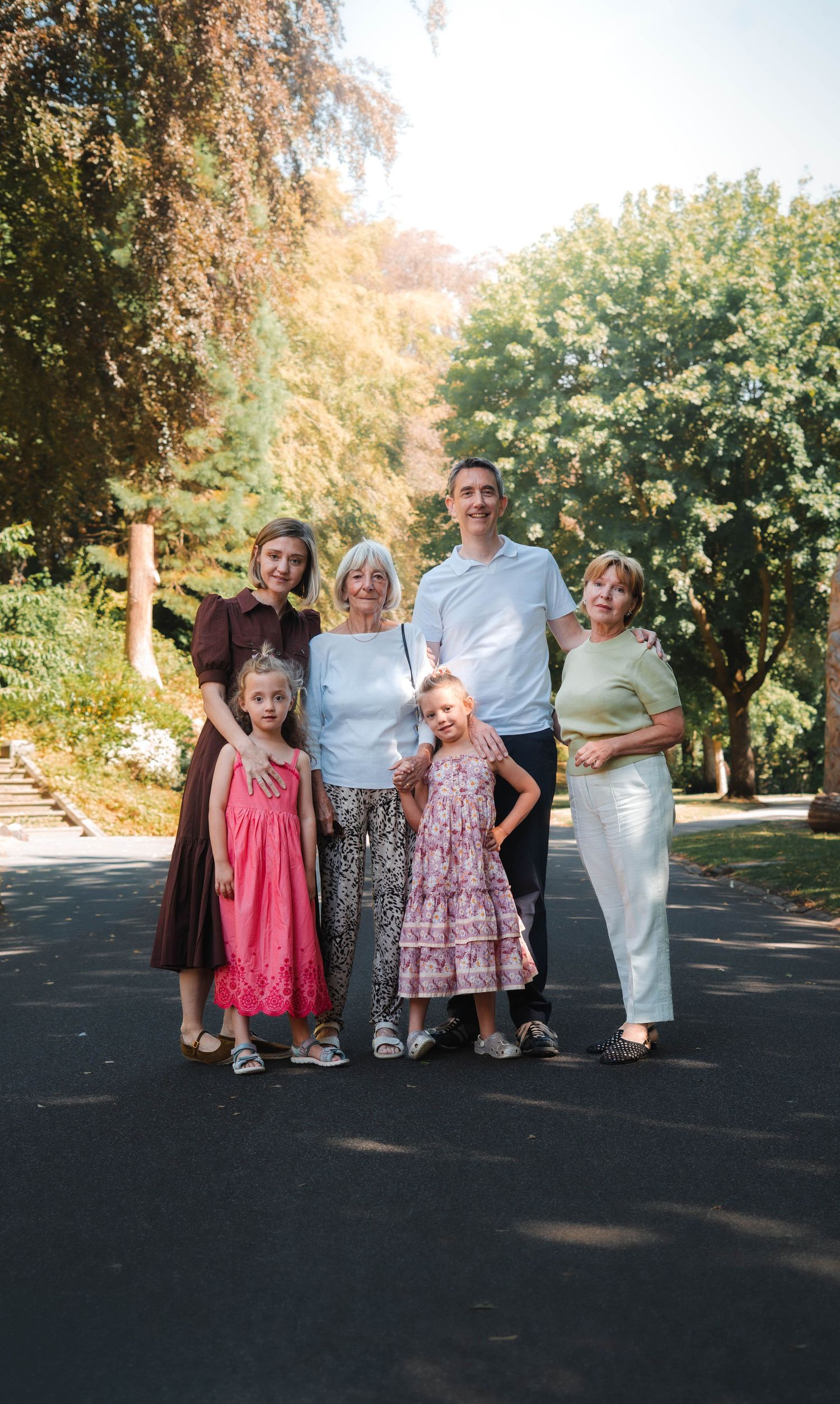 Multi-generational family portrait with two children and four adults posing outdoors on a sunny tree-lined path.