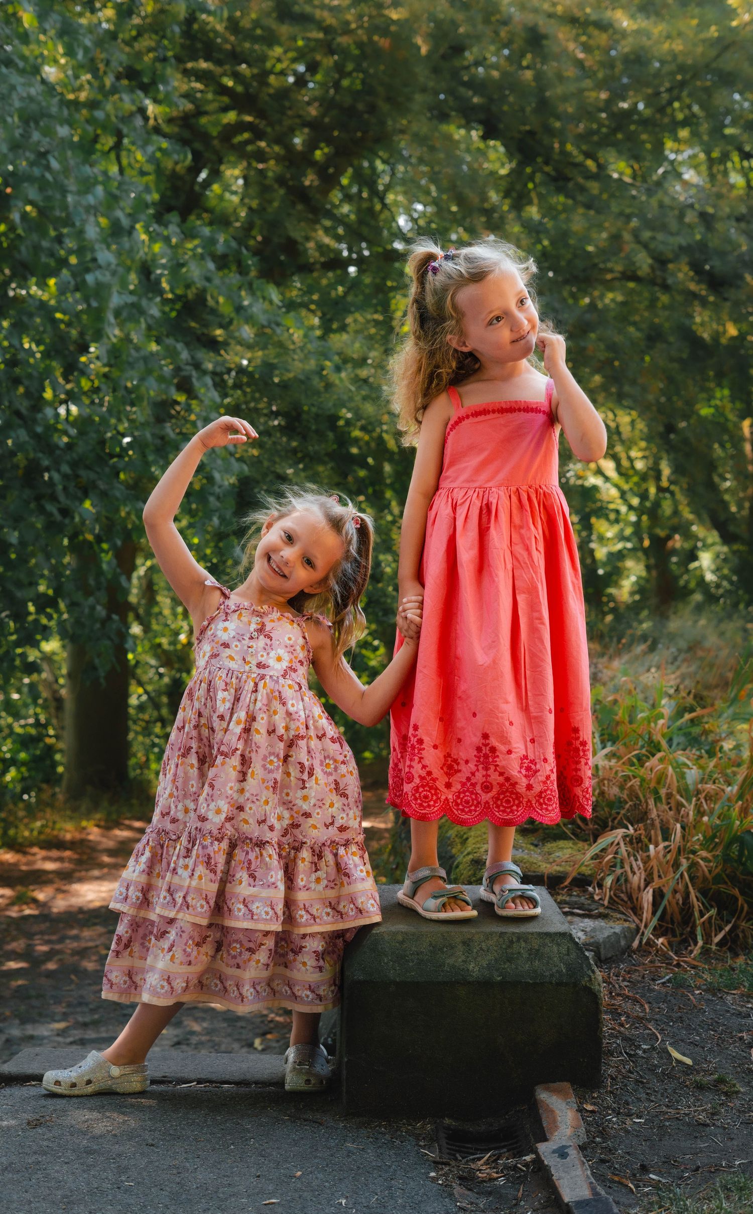 Two young girls in pink and coral summer dresses posing outdoors among green trees.
