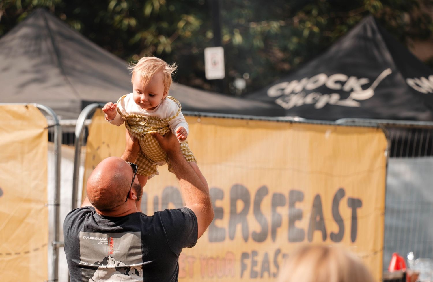 Adult lifting smiling baby in gold outfit high in the air at outdoor festival.