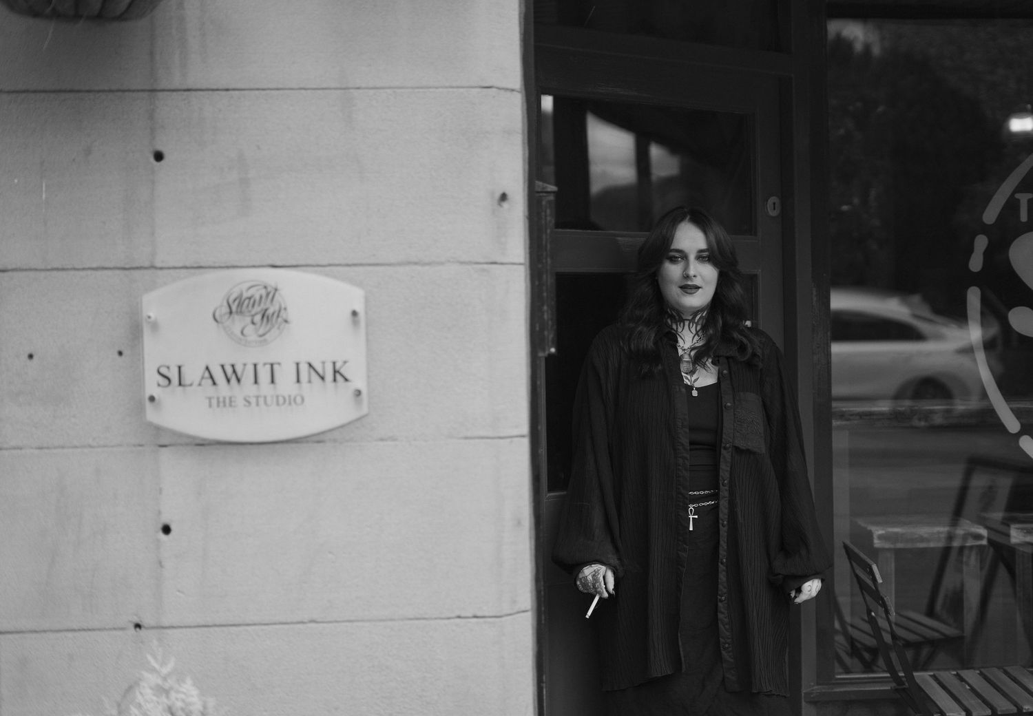 Black and white portrait of a woman standing outside Slawit Ink tattoo studio entrance.