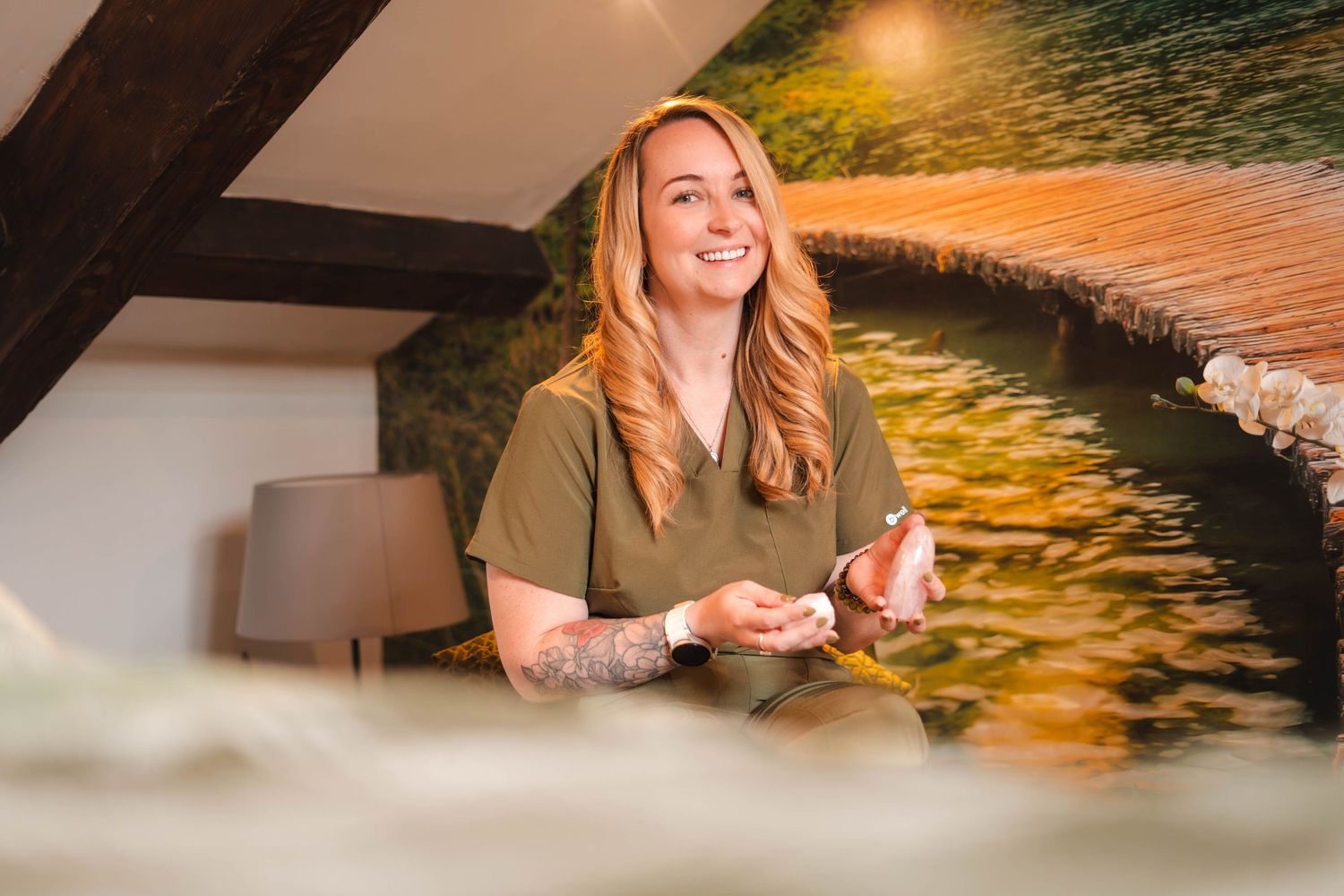 Smiling blonde woman in olive scrubs sitting indoors near nature mural wall.