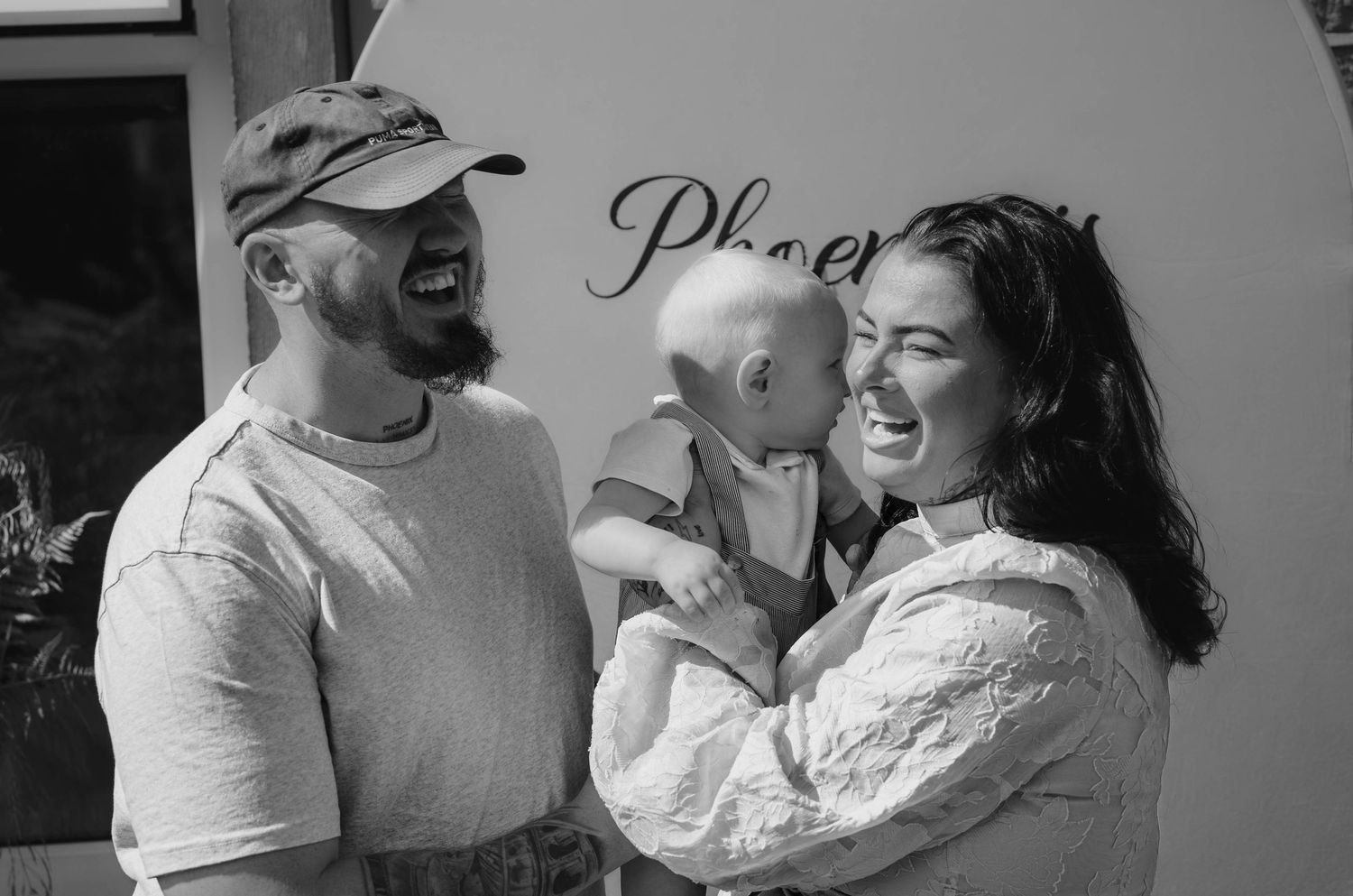 Black and white photo of smiling couple holding laughing baby outdoors near Phoenix sign.