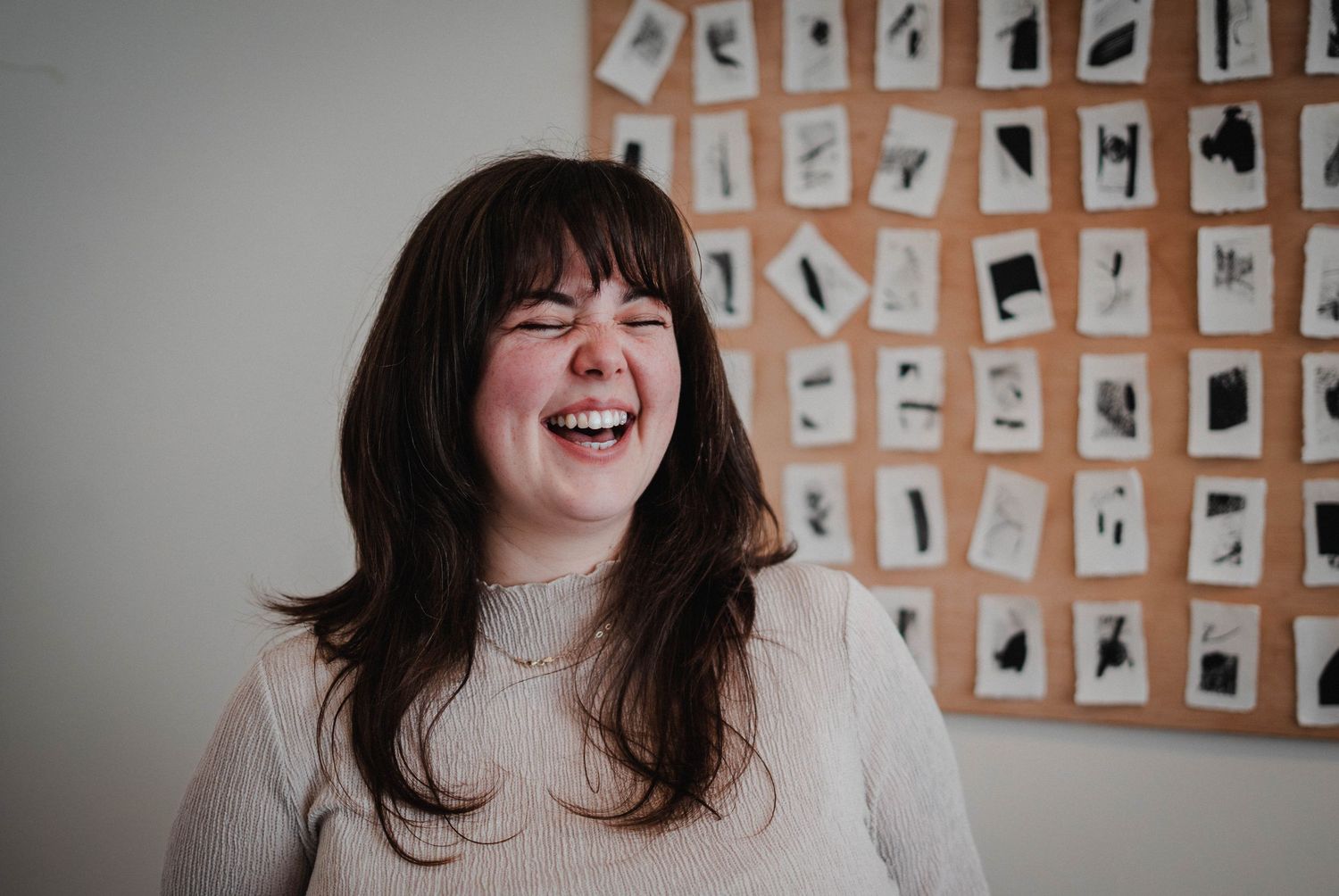 Young woman with dark hair laughing joyfully in front of a cork board displaying black and white photos.