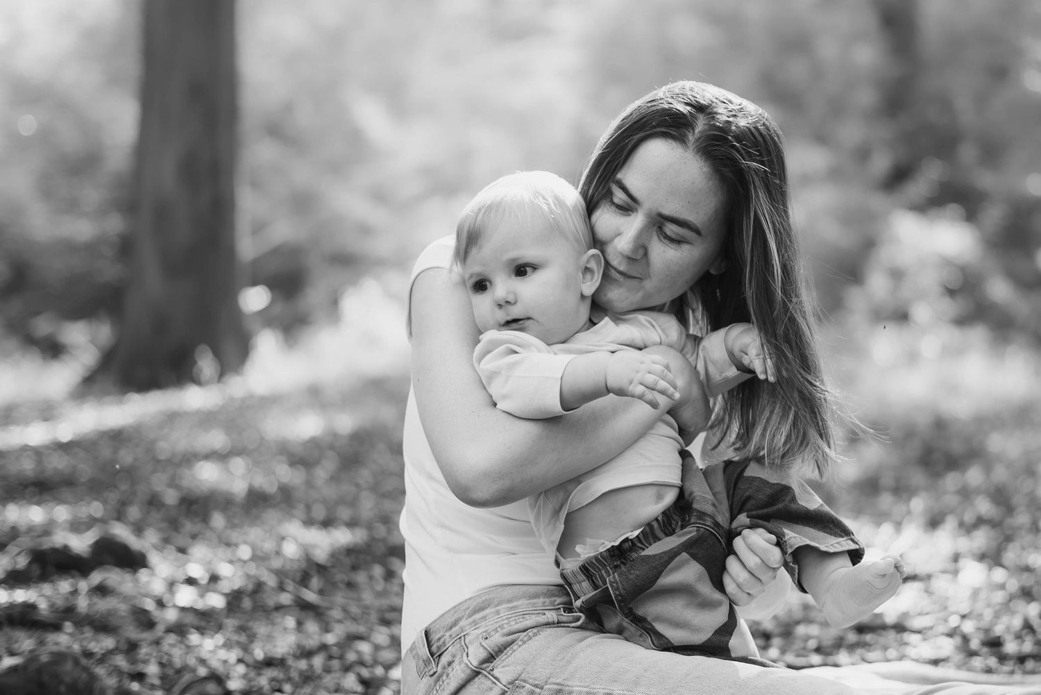 A tender black and white photo of a young mother embracing her baby in a sunlit forest setting.