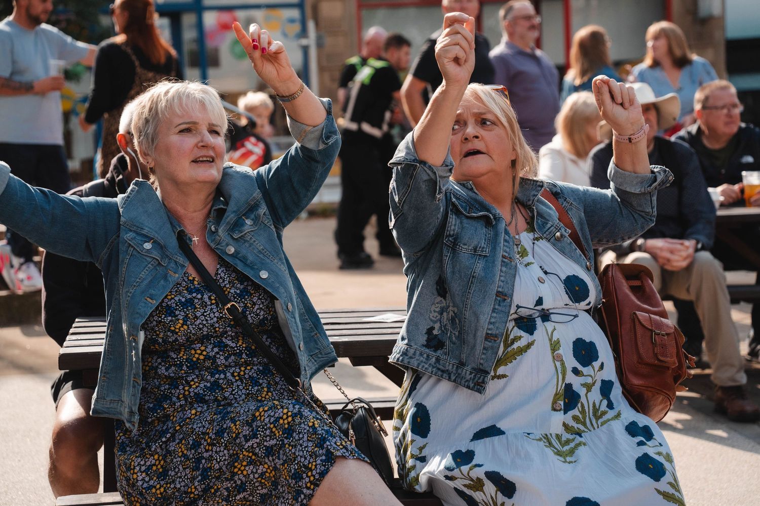 Two older women in denim jackets dancing joyfully on a bench at outdoor event.