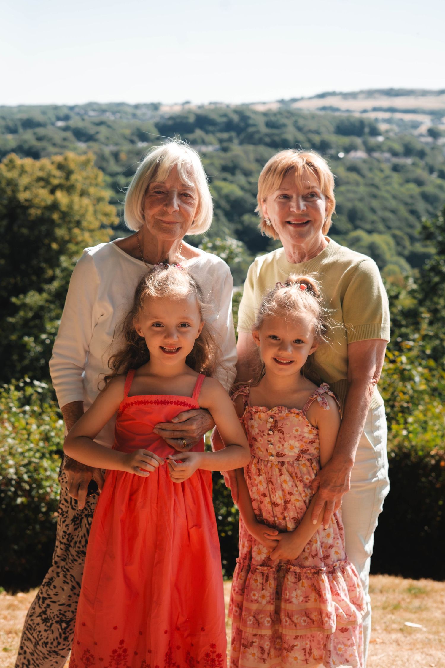 Two grandmothers embrace two young girls in summer dresses with a scenic countryside backdrop.