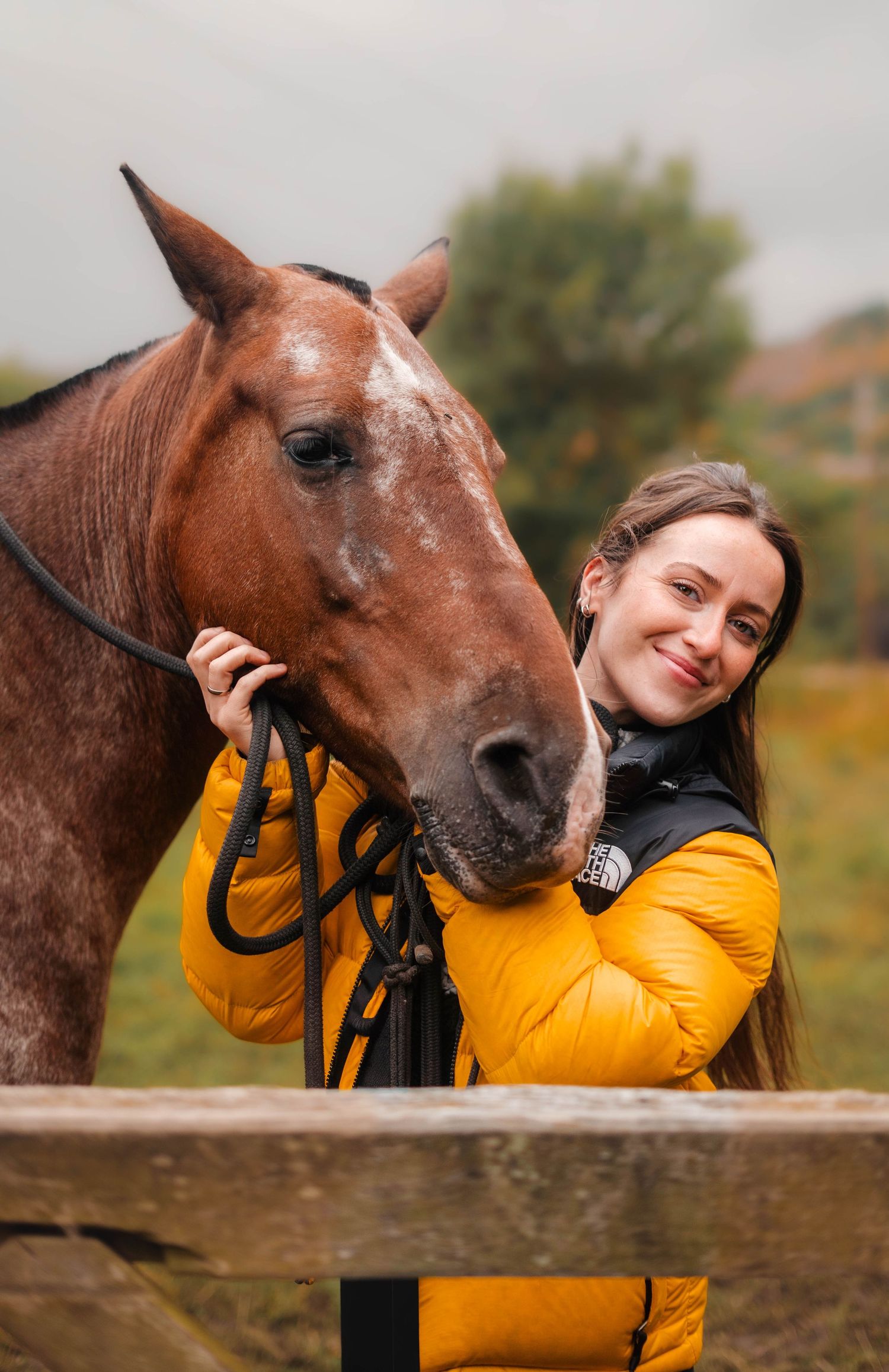Smiling woman in yellow North Face jacket posing with a chestnut horse at a wooden fence.