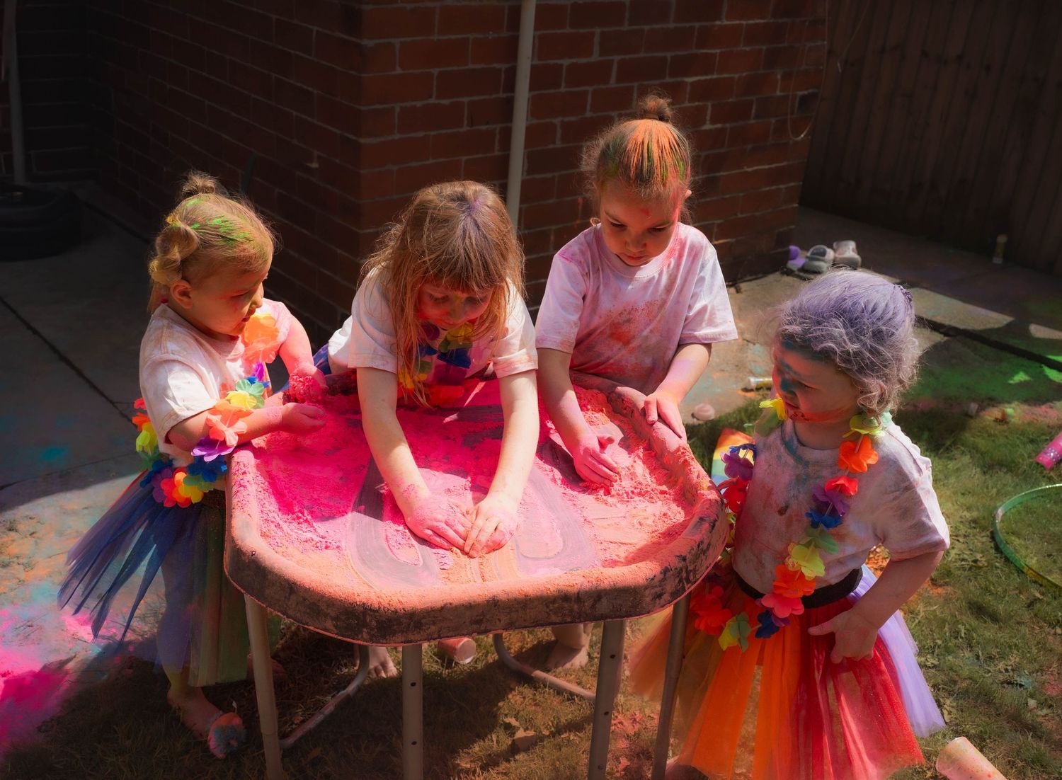 Four children covered in colorful powder paint playing at an outdoor table during a Holi color festival party.