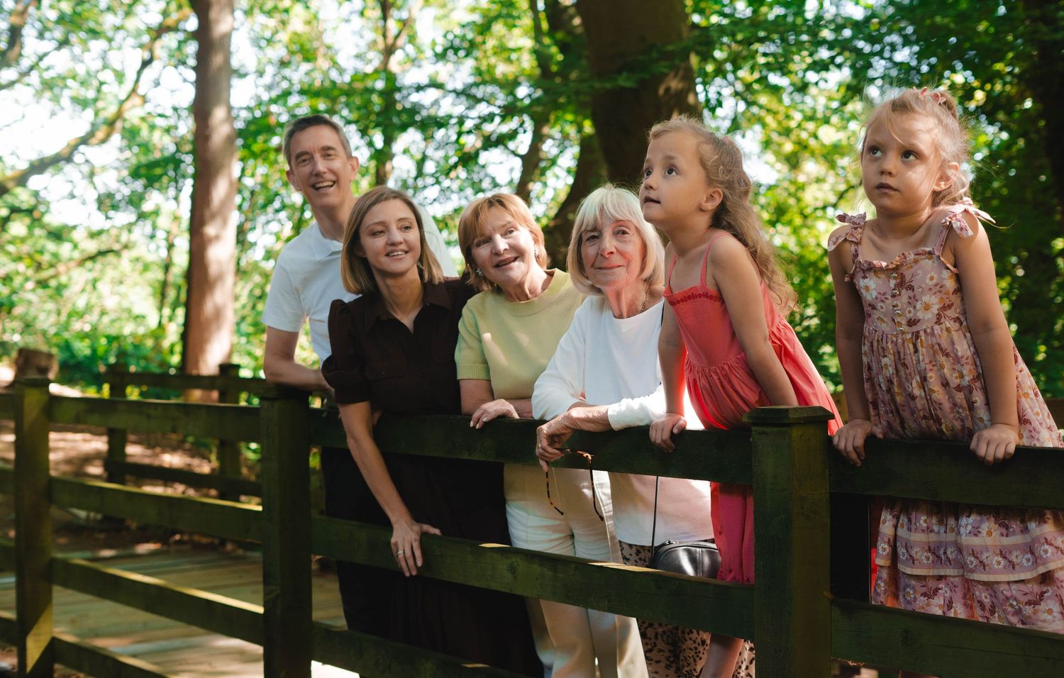 A multi-generational family smiles together leaning on a wooden bridge in a forest.
