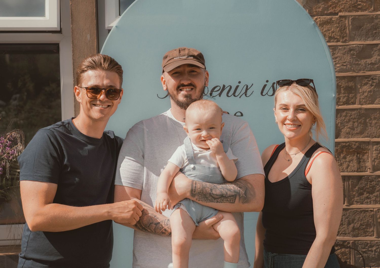 Group of three adults and baby posing outdoors in front of Phoenix Is sign on sunny day.
