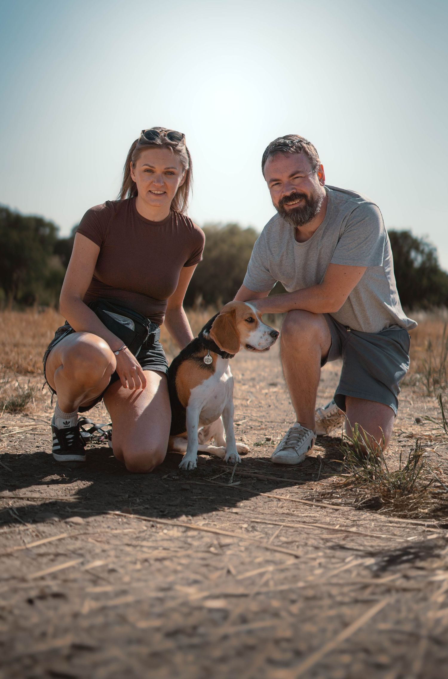 Smiling couple crouching beside a small Beagle dog on a dry dirt path outdoors.