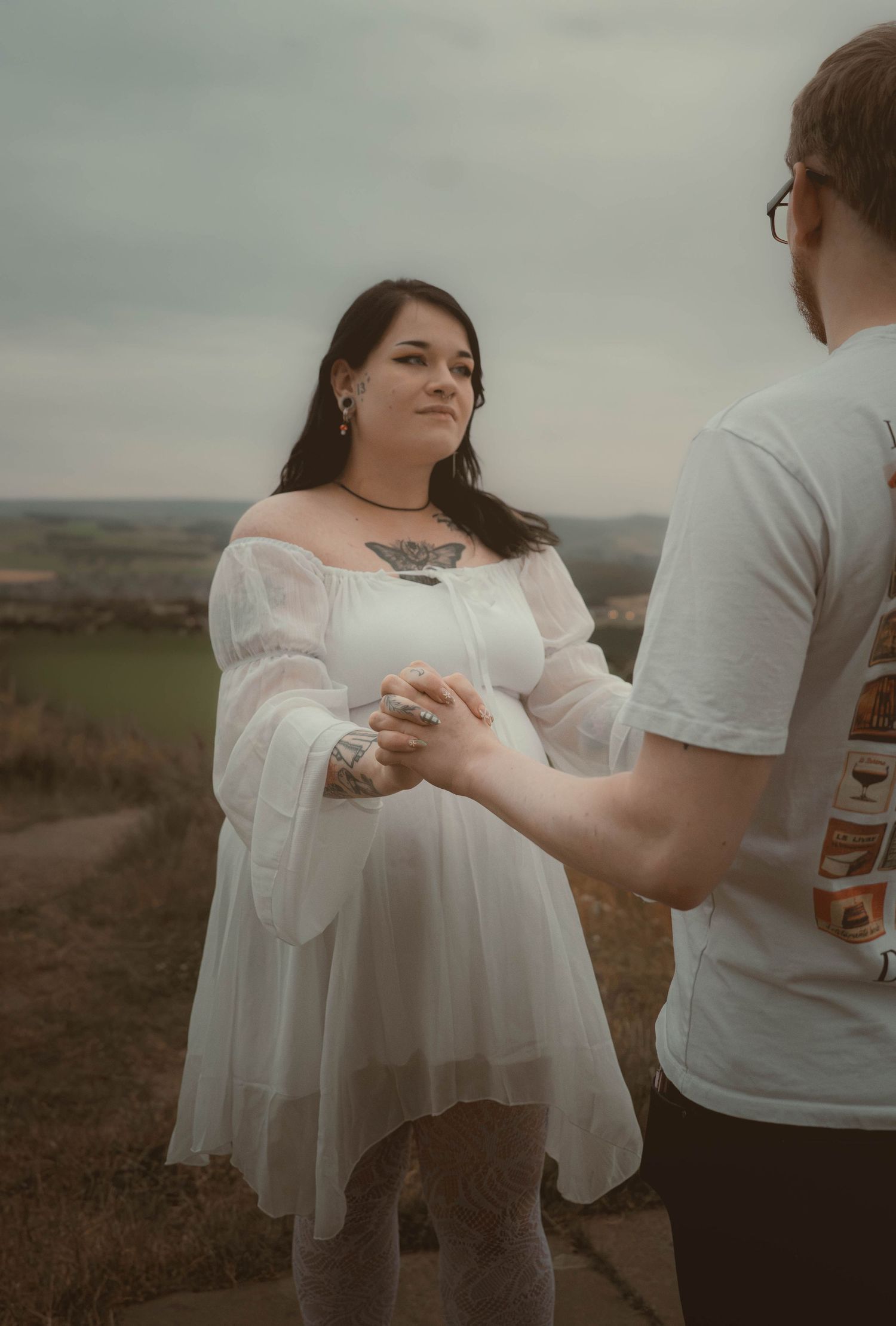 Tattooed pregnant woman in white off-shoulder dress holding hands with partner on scenic hilltop.