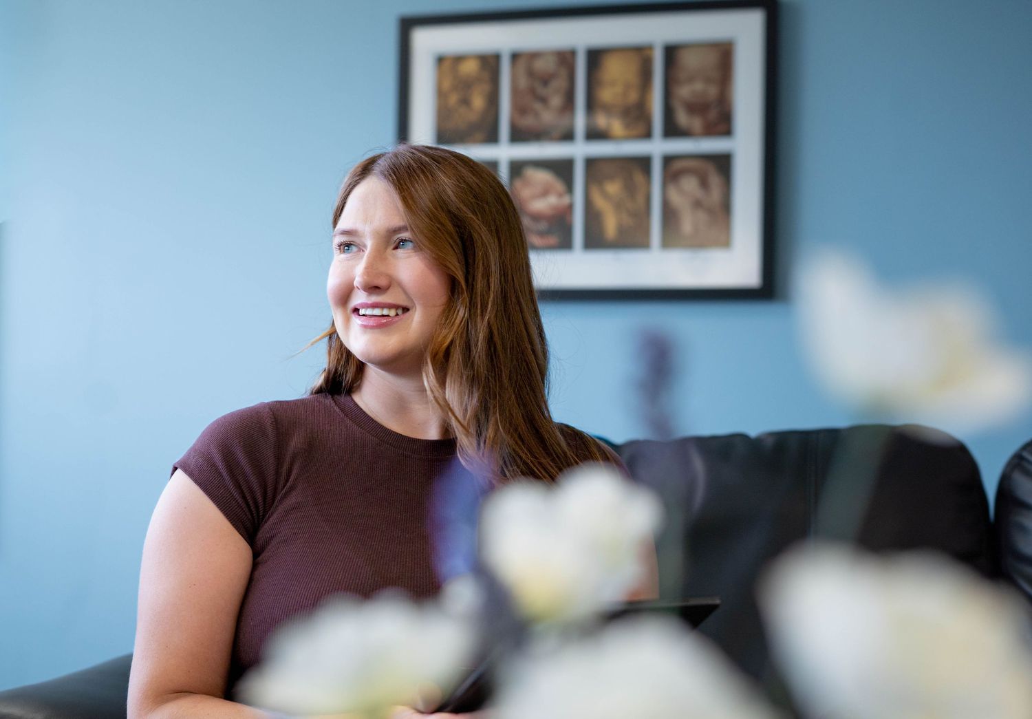Smiling young woman relaxes on a sofa beneath framed ultrasound images on a blue wall.