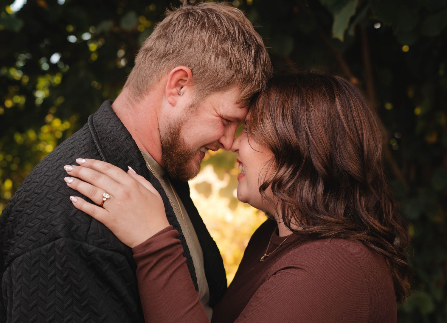 An engaged couple touches foreheads romantically surrounded by golden autumn foliage.