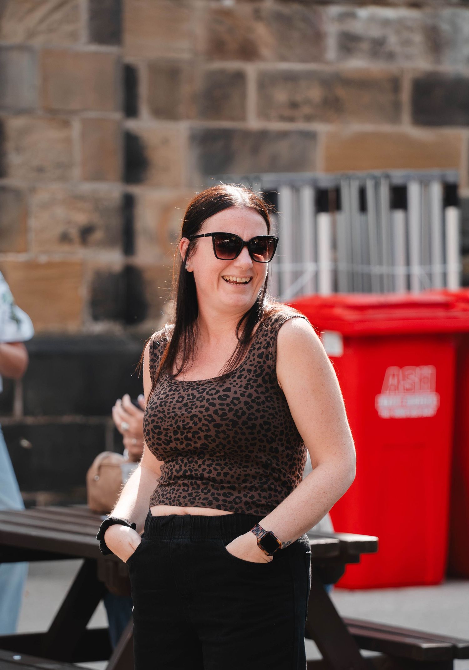 A smiling woman in sunglasses and leopard print top stands outdoors in sunlight.
