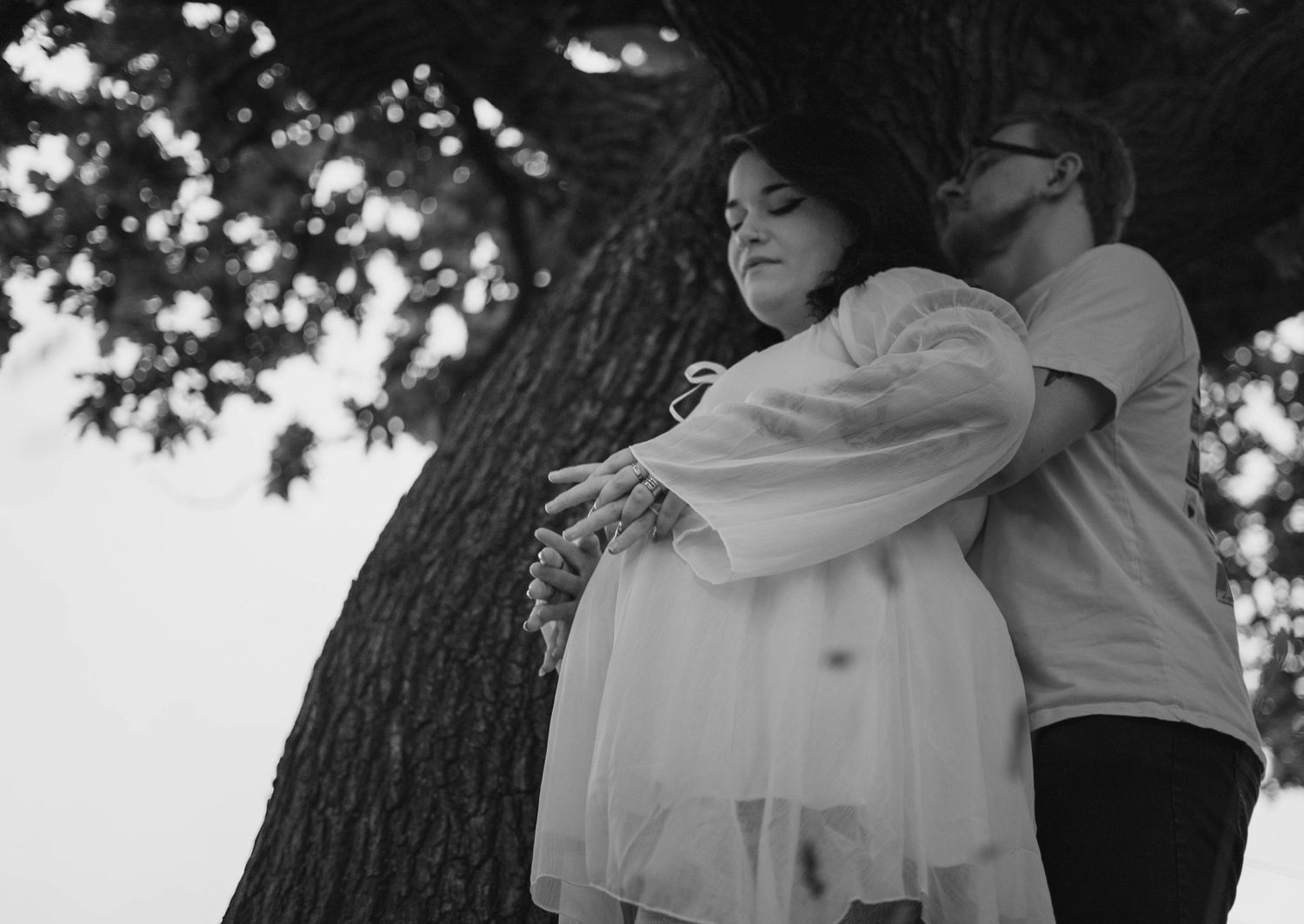 A couple embraces tenderly beneath a large tree in a romantic black and white outdoor portrait.