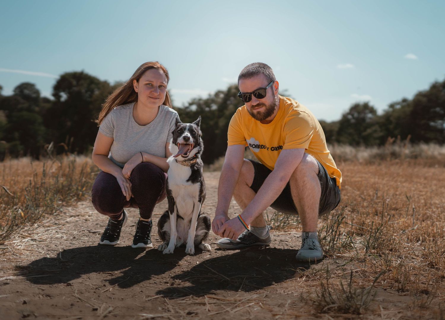 A couple squats beside their black and white Border Collie on a sunny dirt trail.