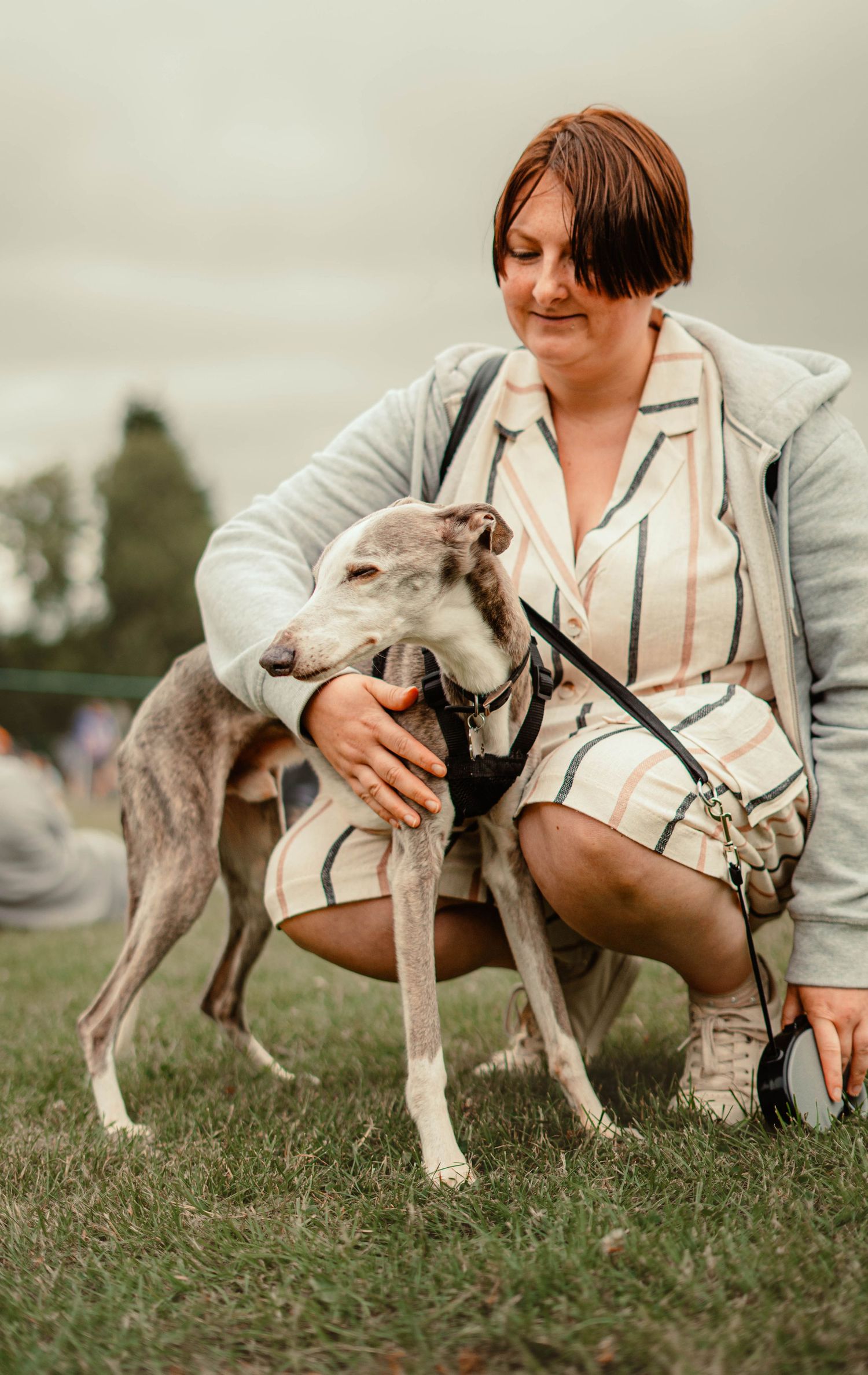 Woman crouching and hugging a grey Greyhound dog on grass at an outdoor event.