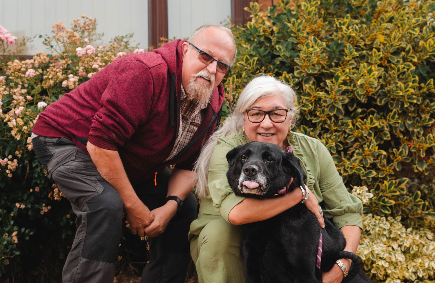 Smiling elderly couple posing outdoors with their black Labrador dog beside colorful garden shrubs.