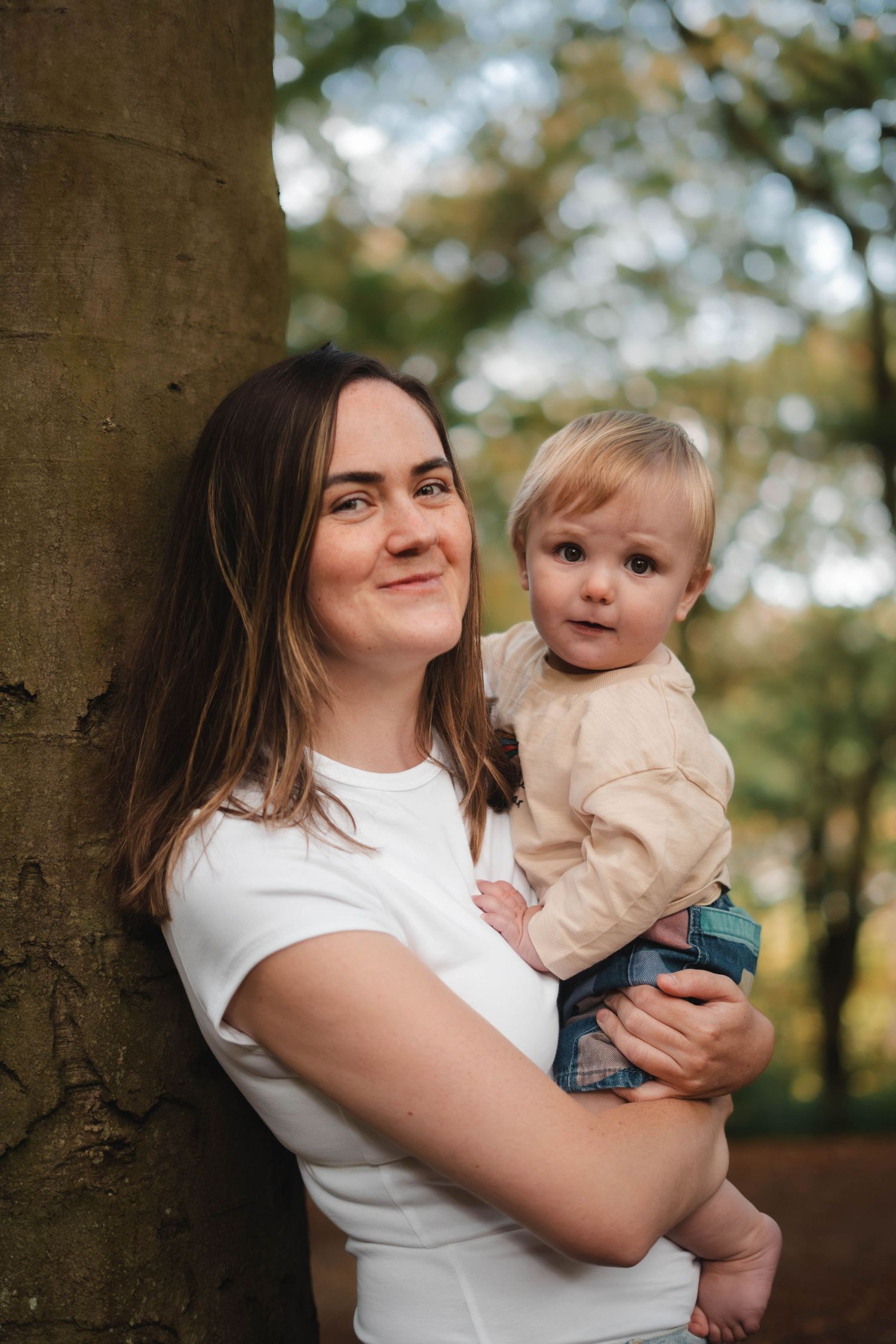 A young mother leans against a tree holding her blonde baby in an autumn park.