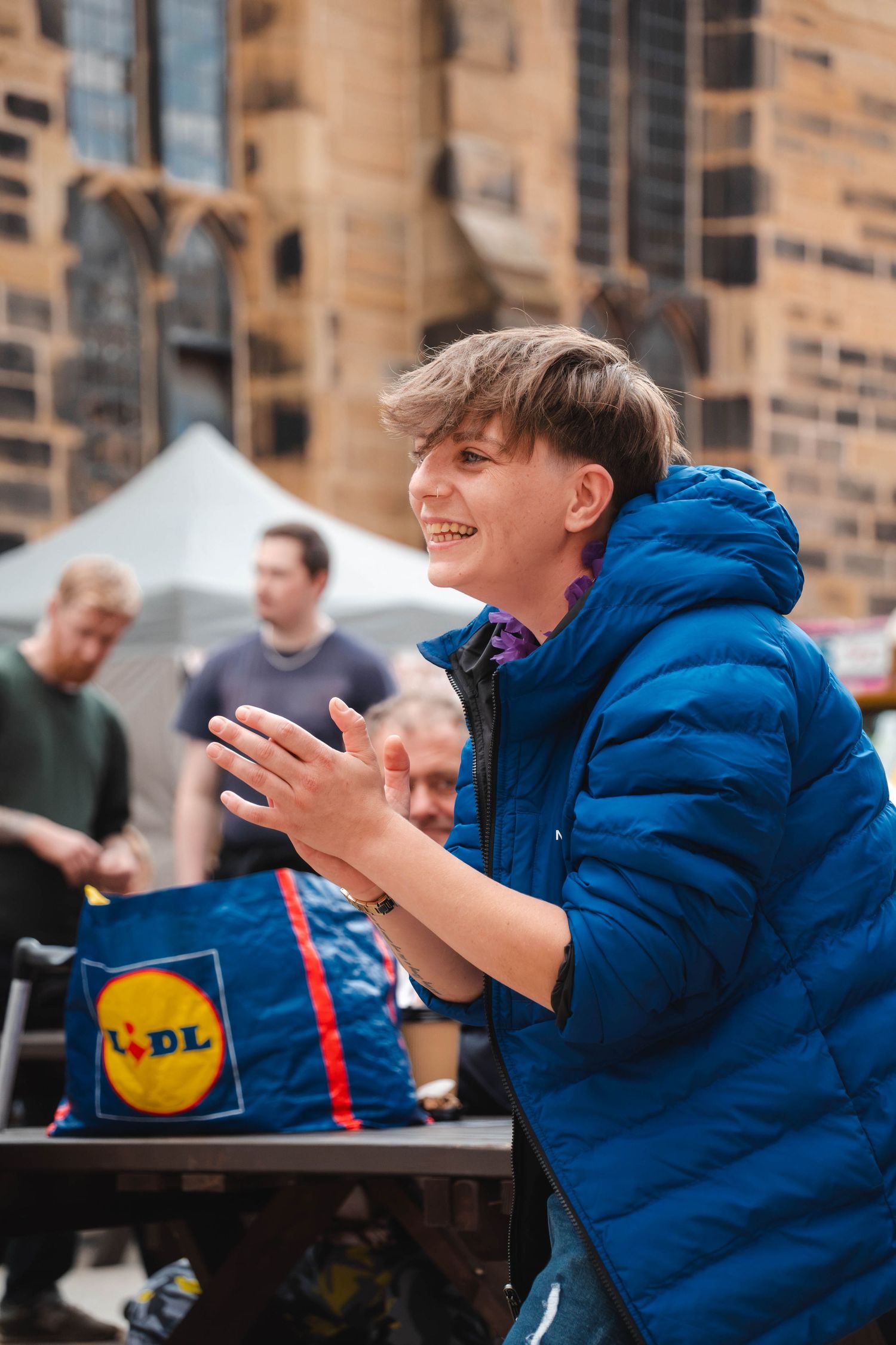 Happy teenager in blue puffer jacket clapping hands at outdoor market near Lidl bag.
