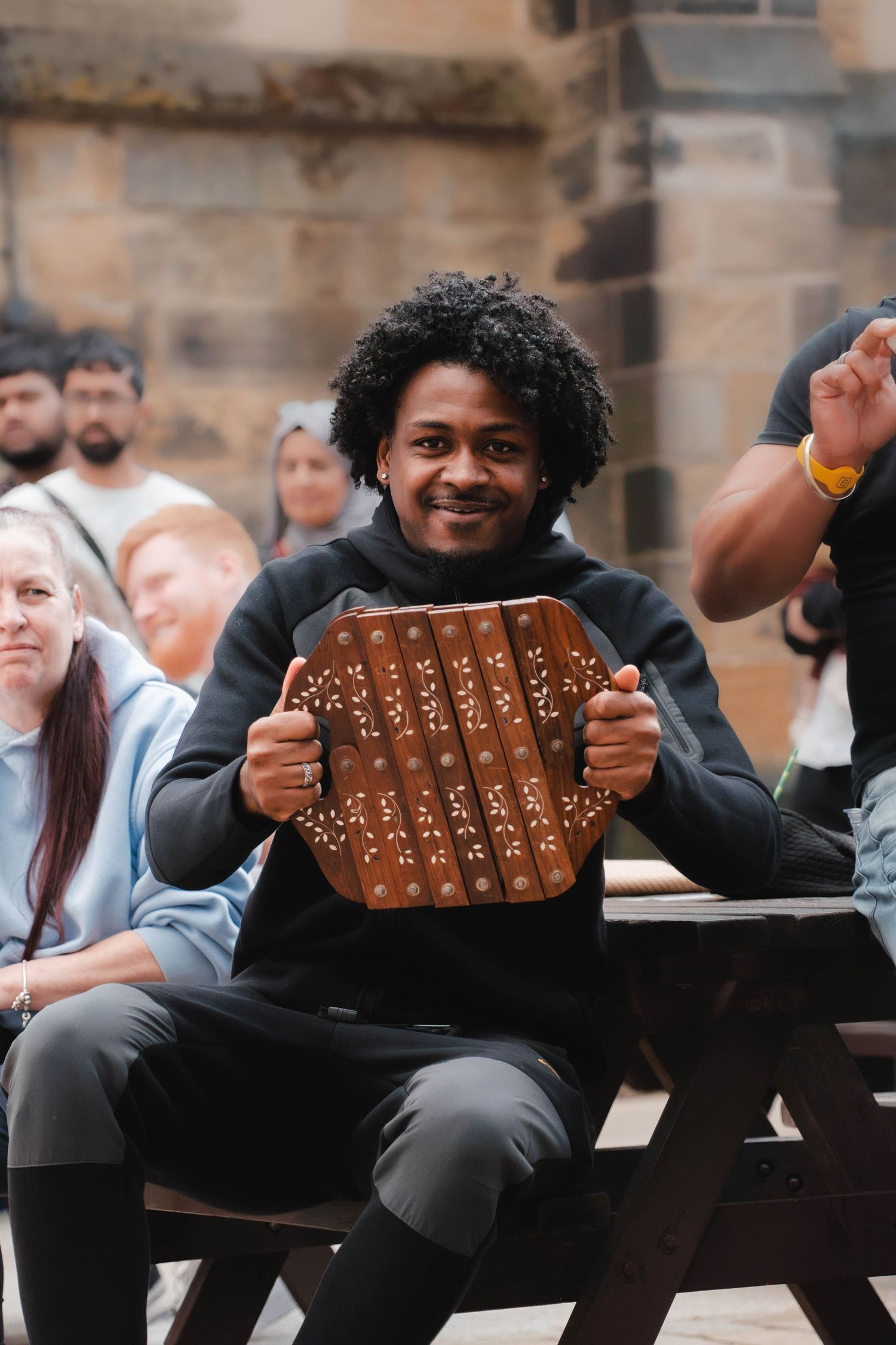 Smiling man holding a wooden kalimba instrument while sitting at a picnic table in a crowd.