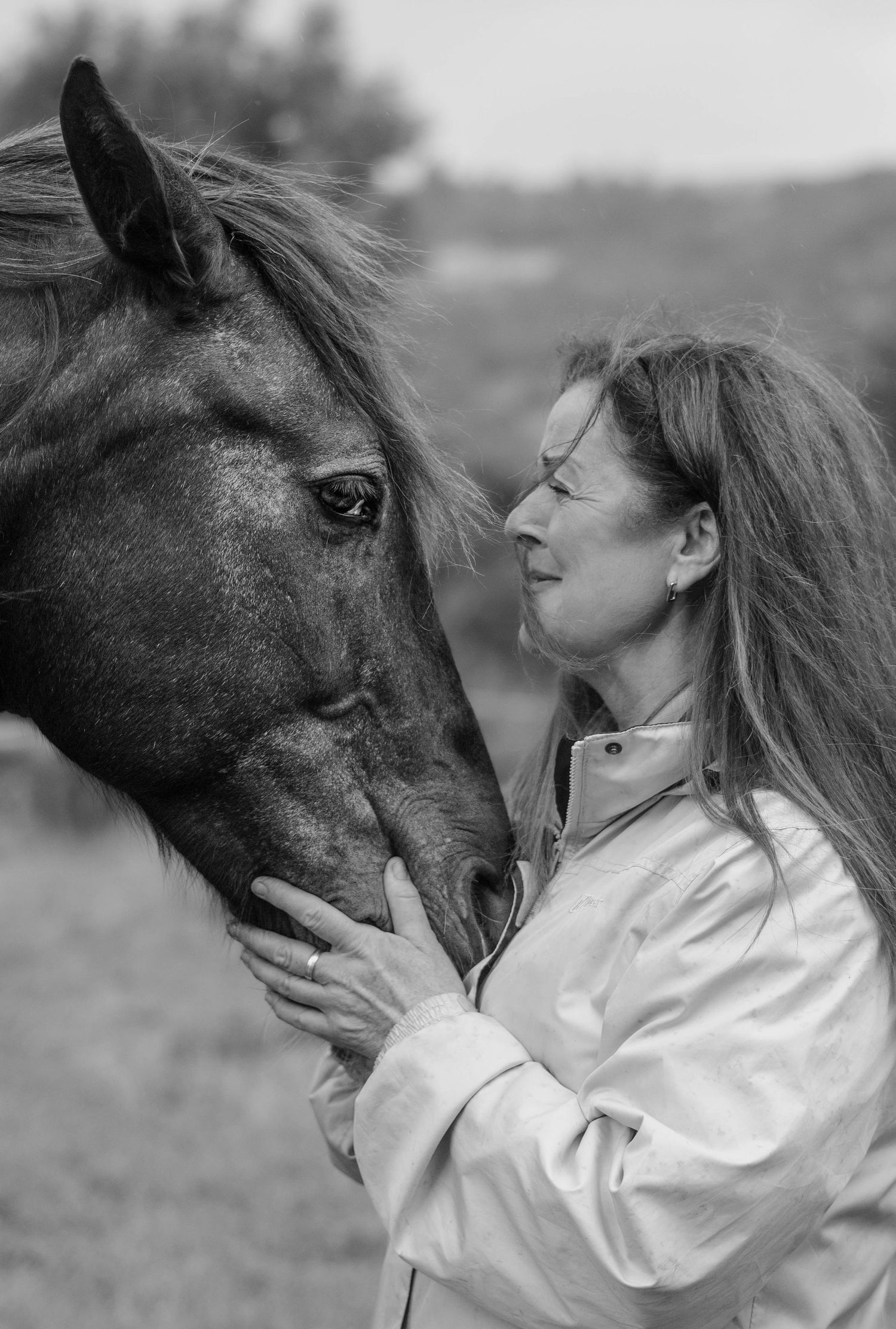 Black and white photo of a woman gently touching foreheads with a horse outdoors.