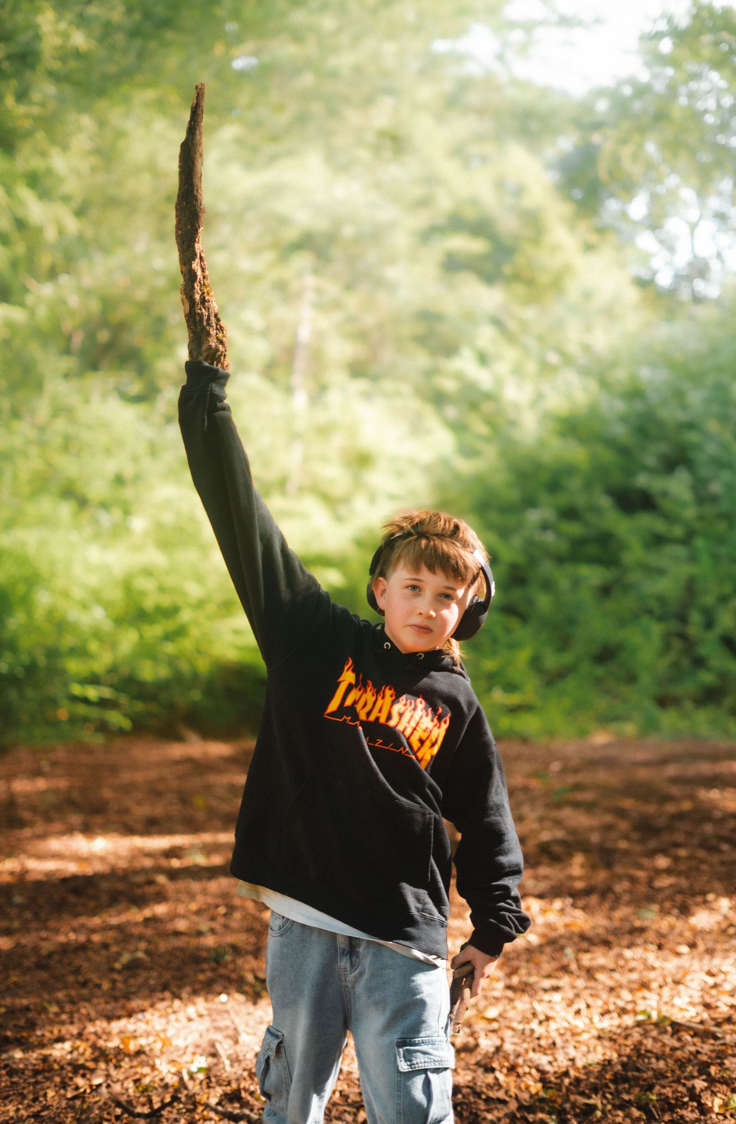 A boy in a Thrasher hoodie raises a stick triumphantly in a sunlit autumn forest.