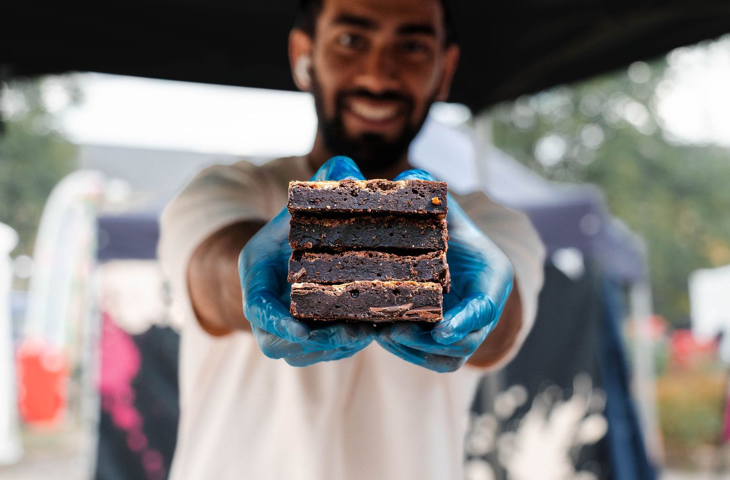 Man in blue gloves holding layered chocolate brownie stack at outdoor food festival.