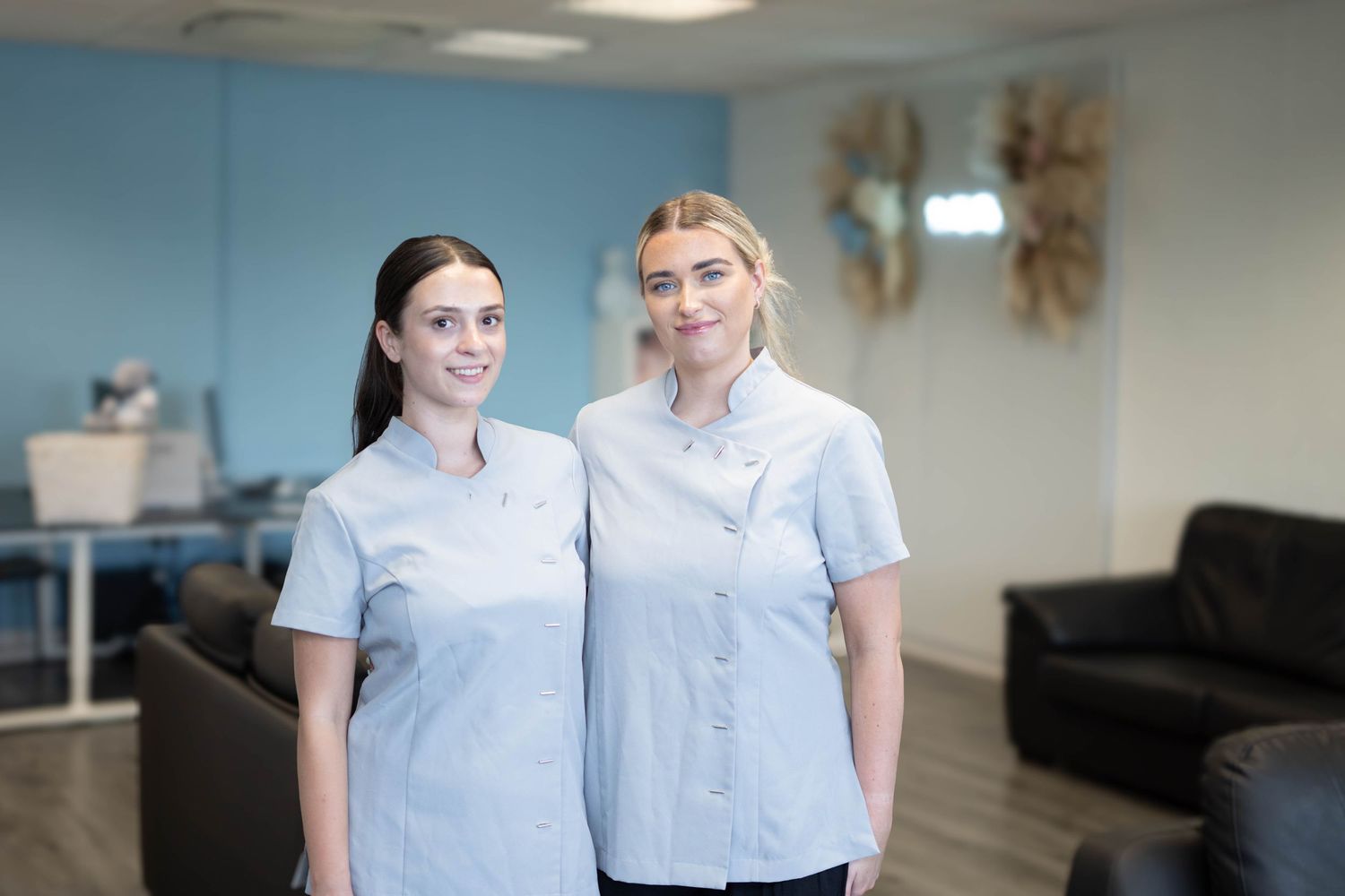 Two female healthcare professionals in light blue uniforms smile together in a medical clinic.