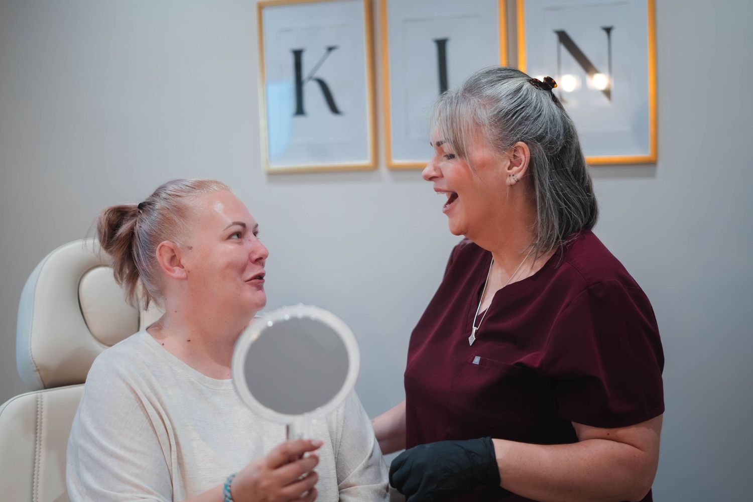 Beauty therapist in dark scrubs showing client mirror reflection results in treatment room.