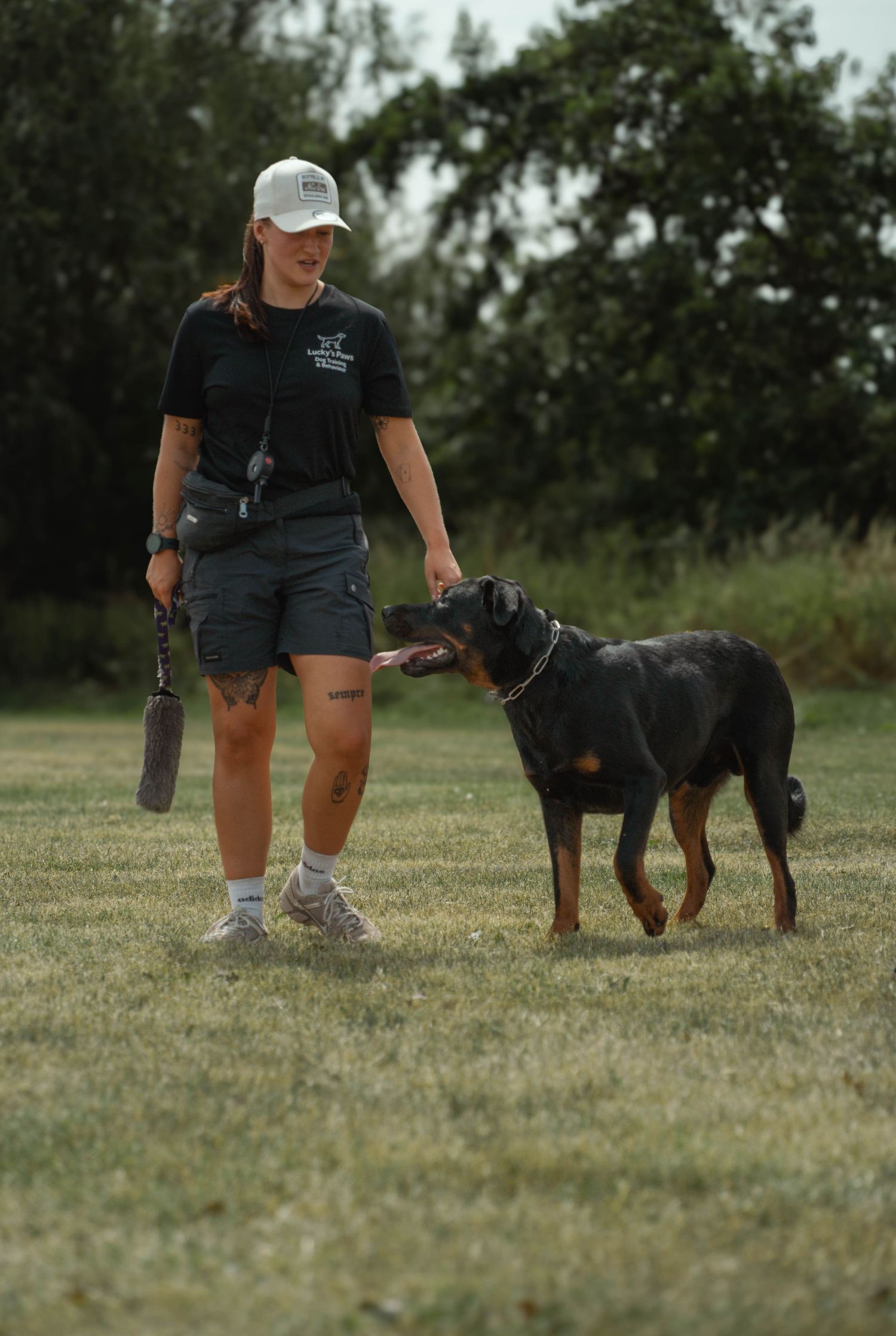 Smiling female dog trainer in white cap walking a Rottweiler on a leash across a grassy field.