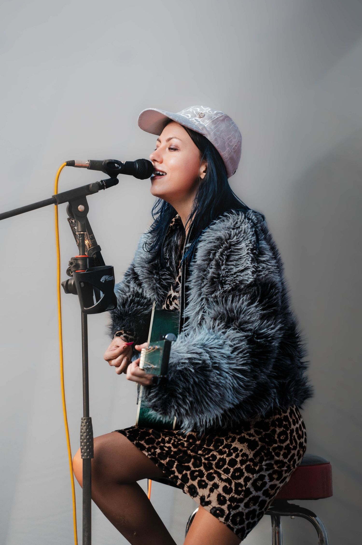 Female singer performing acoustic guitar while wearing fur coat and cap at microphone.