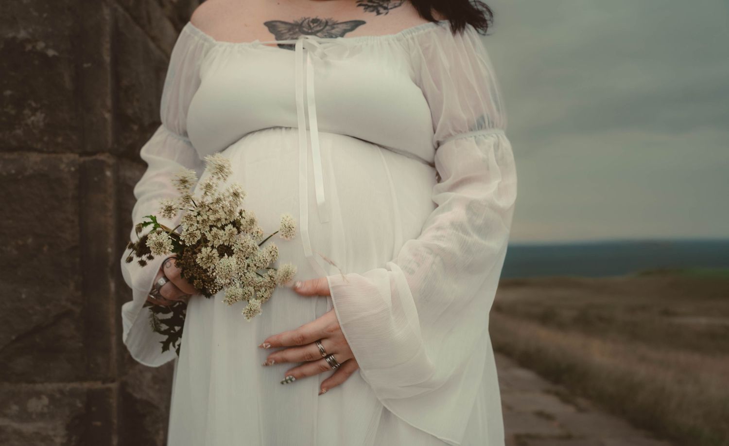 A pregnant woman in a sheer white maternity dress holds a bouquet of wildflowers, gently cradling her baby bump.