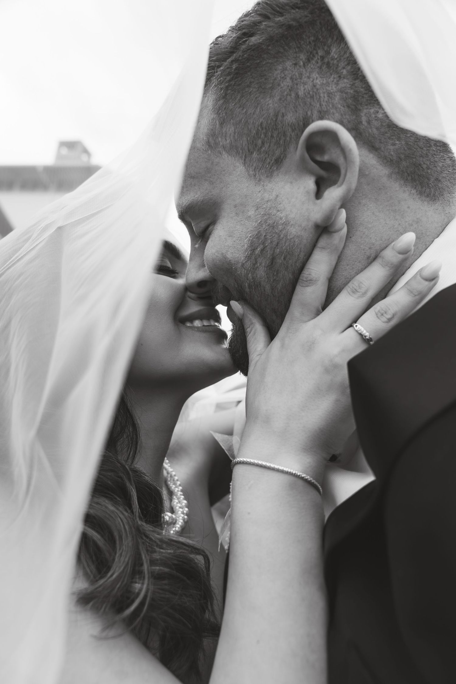 Black and white photo of bride and groom sharing a romantic kiss under a wedding veil, with rings visible.