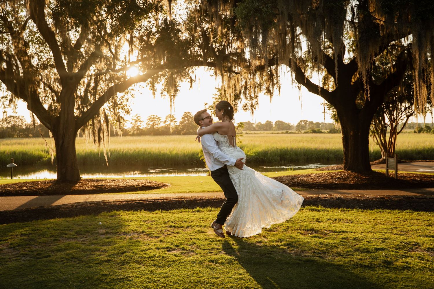 Groom lifting bride under Spanish moss-draped oak trees at golden sunset by a marsh.
