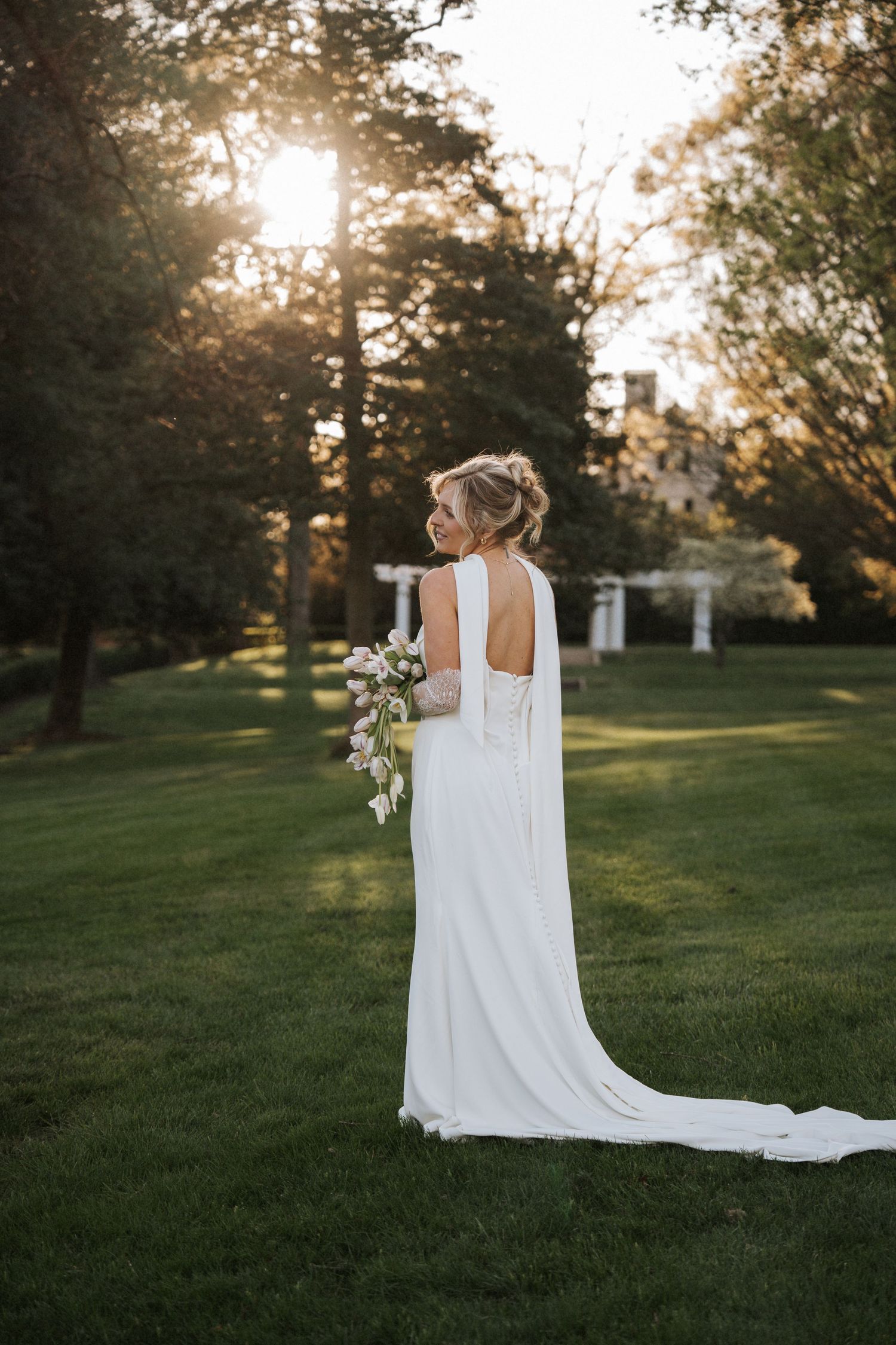 Bride in elegant white backless gown with cape train holds bouquet at golden hour in lush garden.