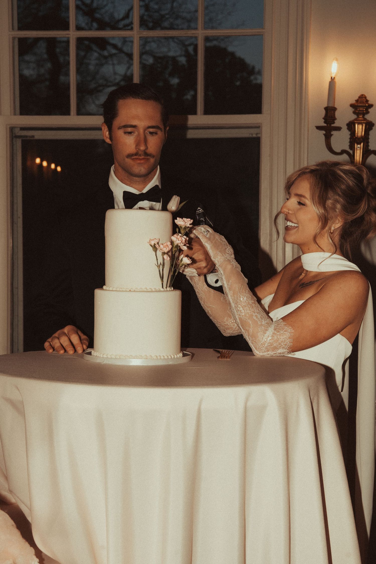 Bride and groom cutting a three-tier white wedding cake decorated with flowers at an elegant evening reception.