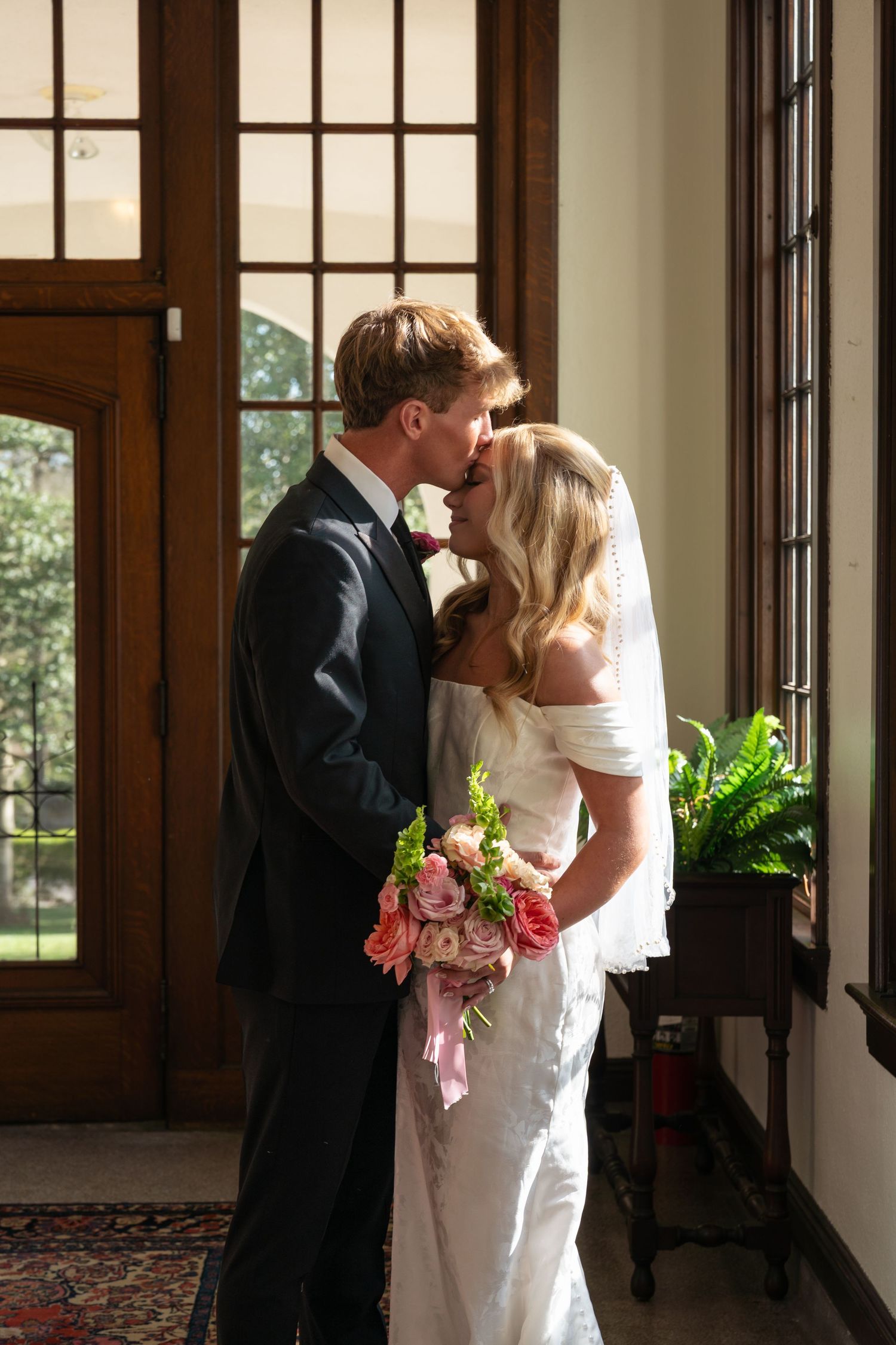 Bride and groom sharing a kiss in elegant venue, bride holding pink bouquet, wearing white gown with veil.