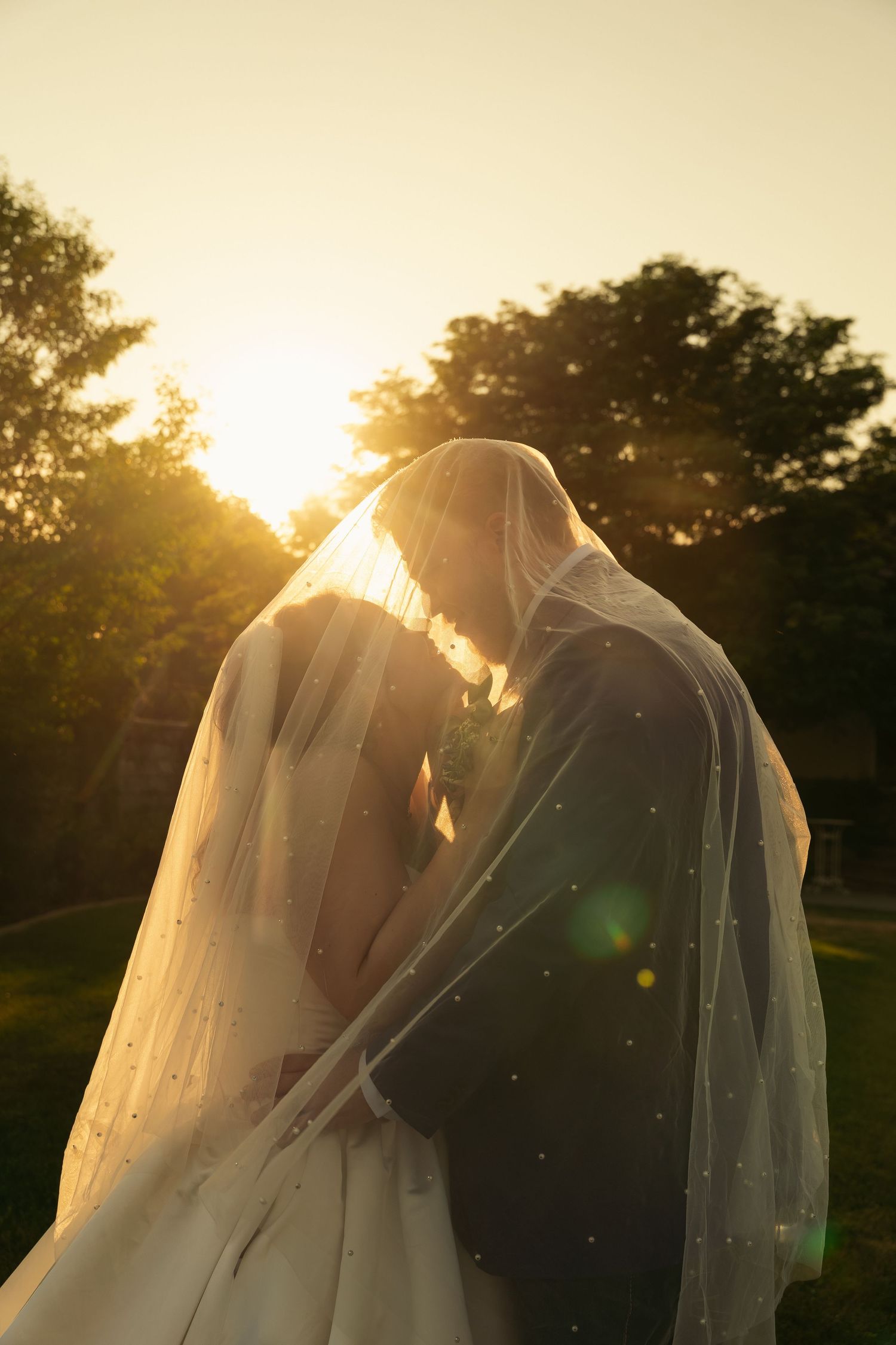 Bride and groom sharing an intimate moment under a veil at golden hour sunset in a lush garden.
