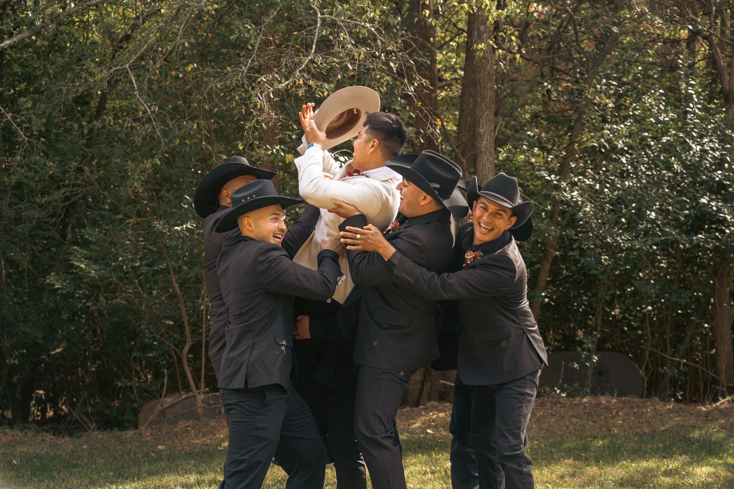 Group of men in black suits and cowboy hats celebrating outdoors, lifting one man in a joyful embrace near trees.