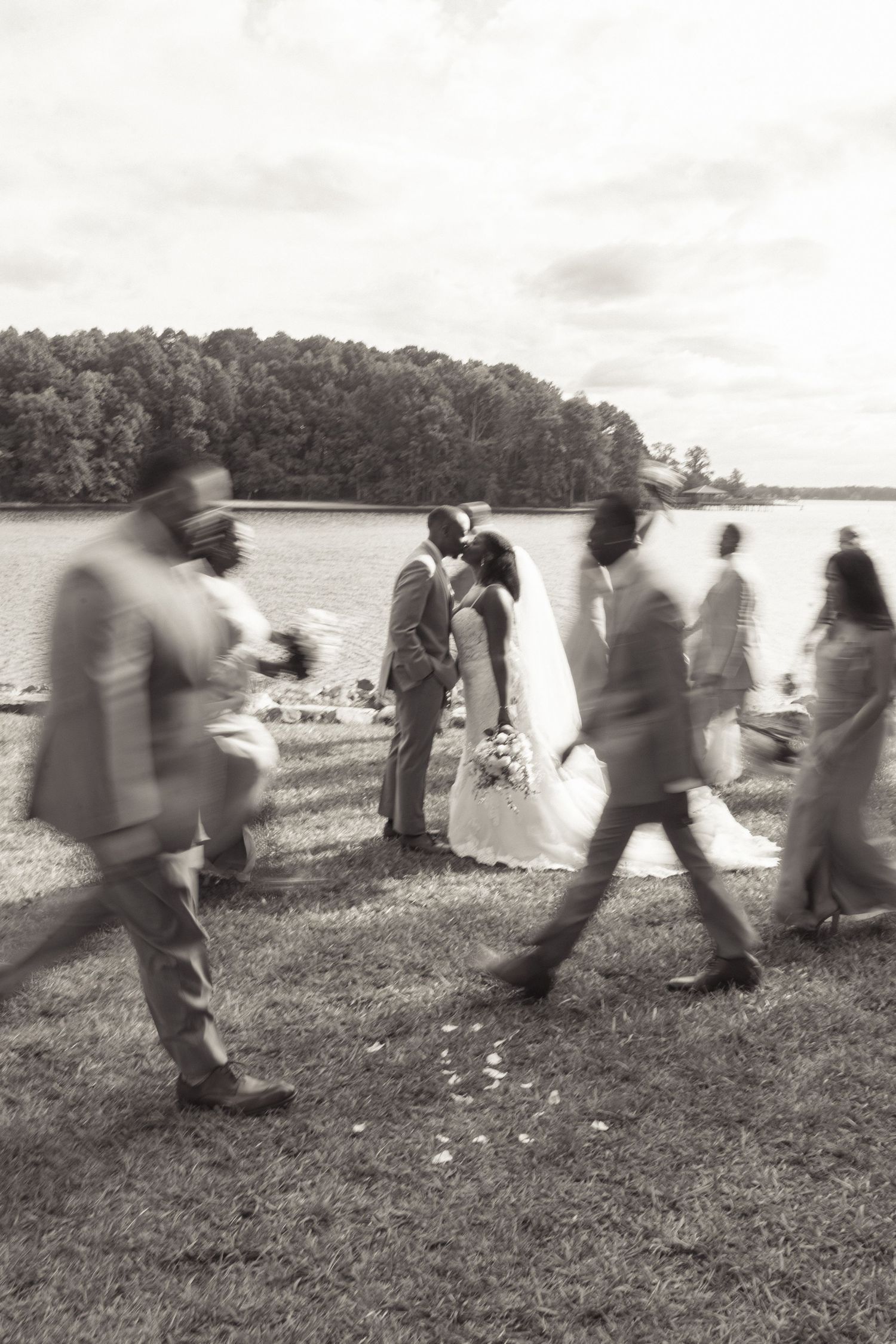 Black and white photo of bride and groom kissing outdoors by a lake while wedding party walks past.