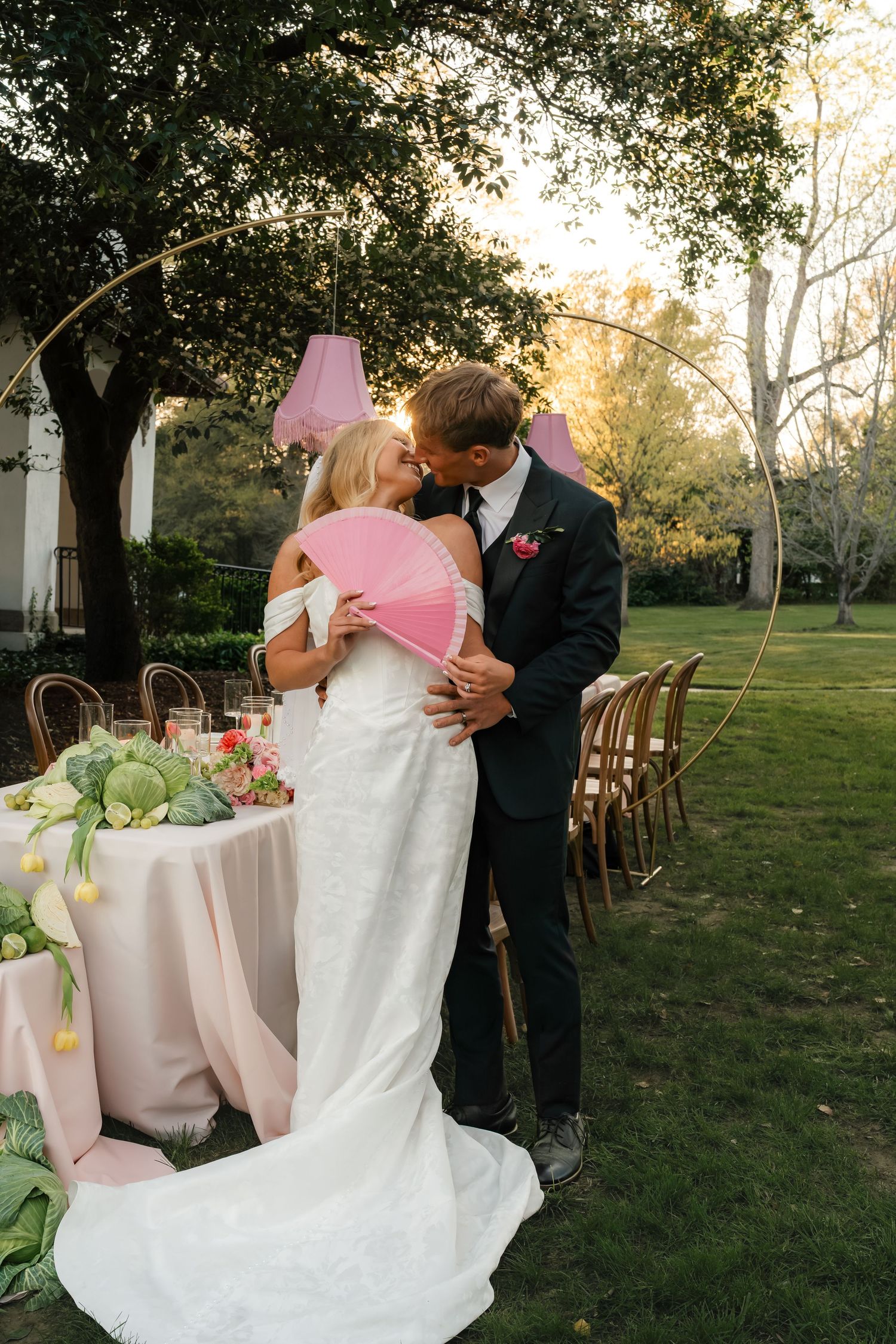 Bride and groom share a kiss at outdoor garden wedding reception, bride holding pink fan near floral tablescape at sunset.