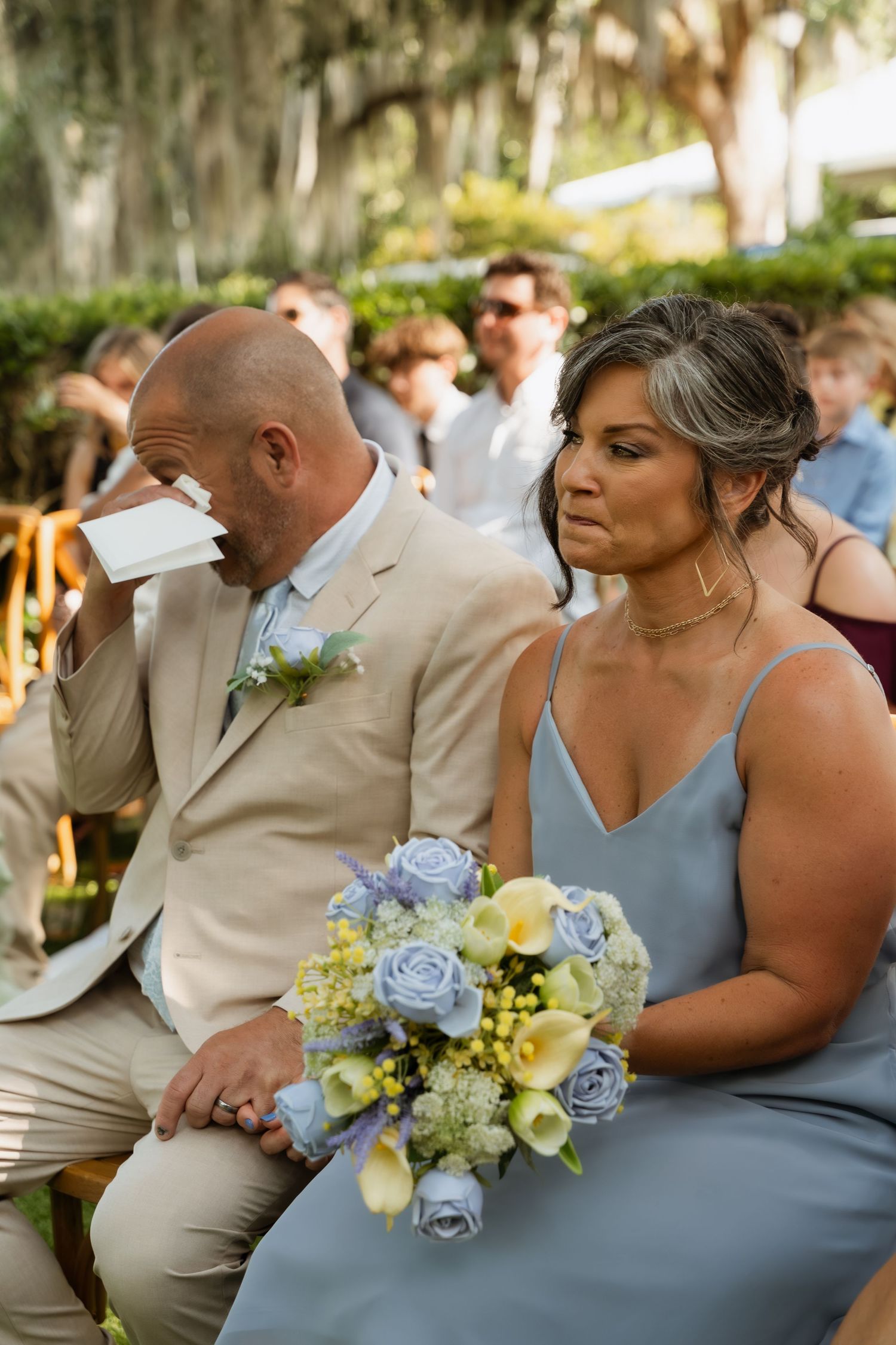 Emotional wedding guest in tan suit dabbing tears beside bridesmaid in blue dress holding floral bouquet at outdoor ceremony.