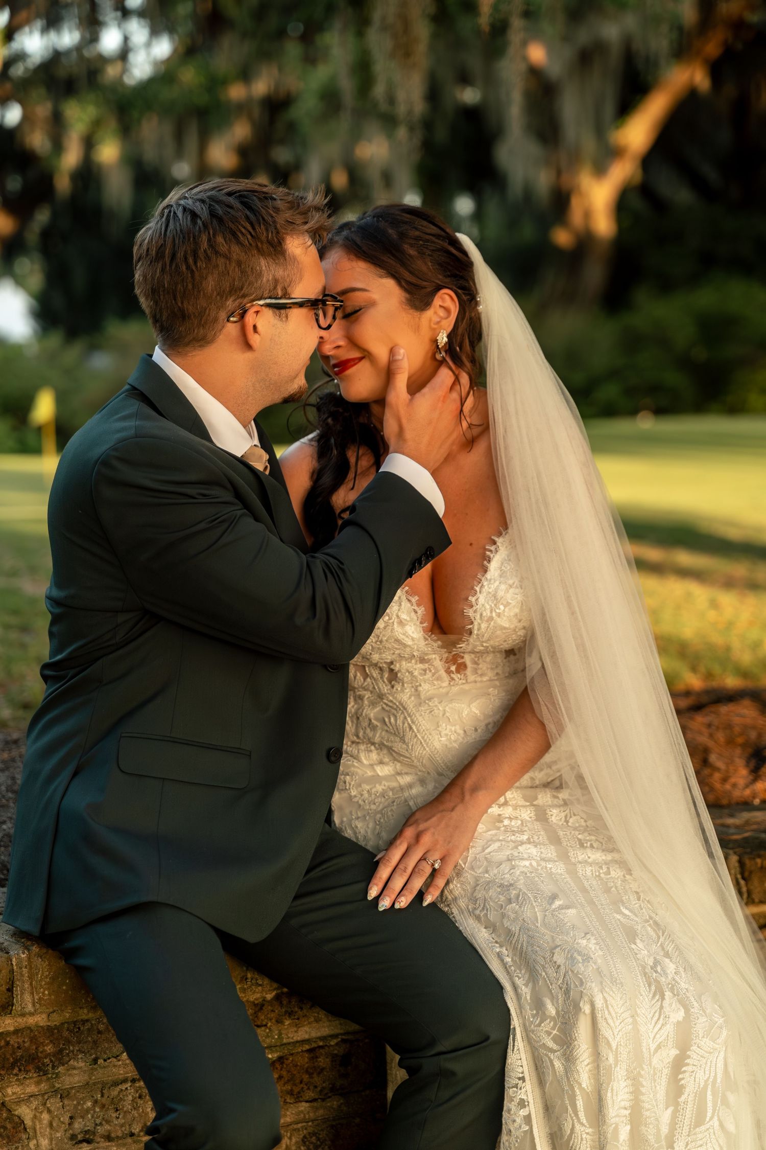 Bride and groom share a romantic moment at golden hour, sitting outdoors amid moss-draped trees.