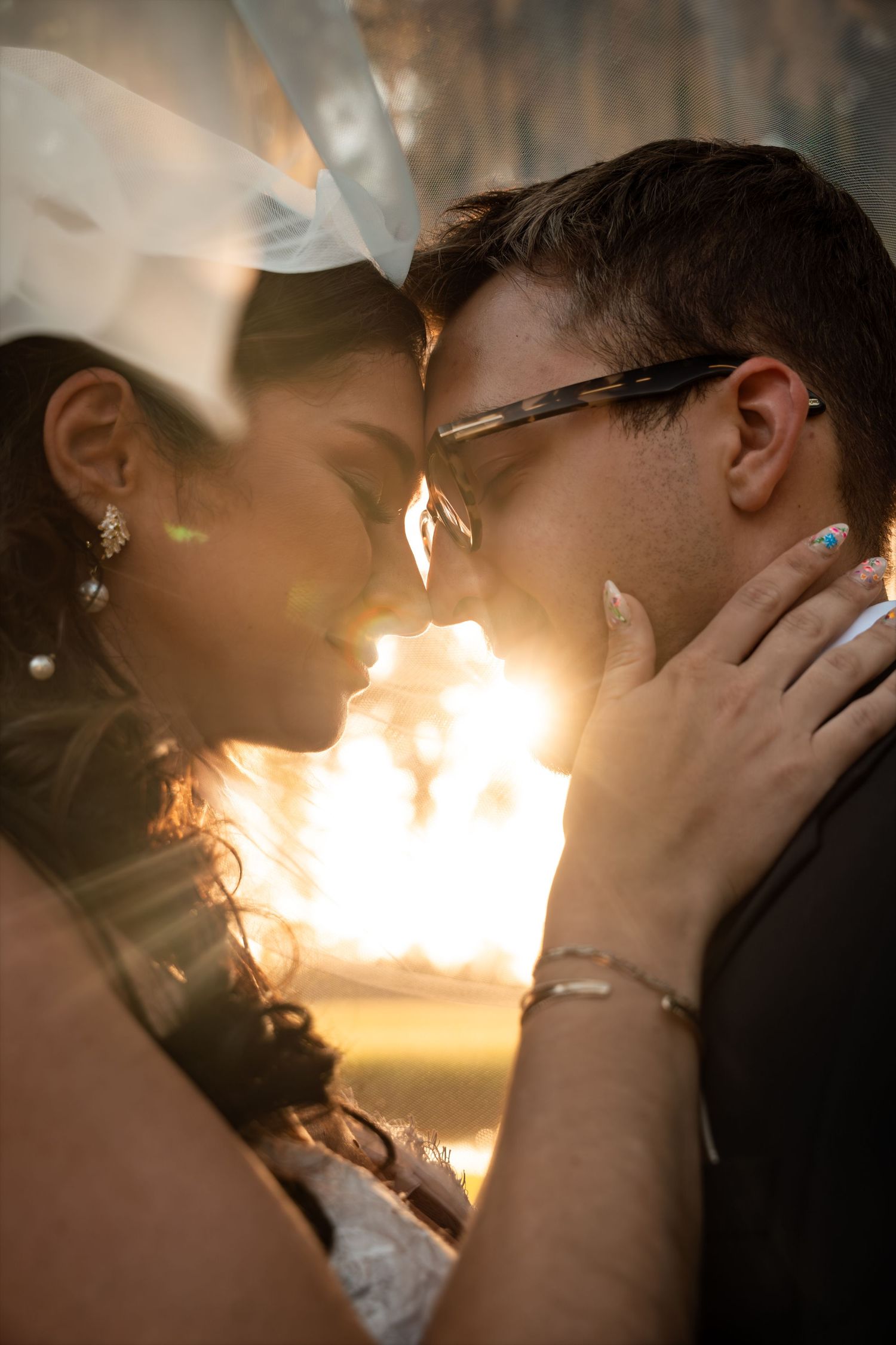 Bride and groom share a romantic moment under a veil with golden sunlight glowing behind them.
