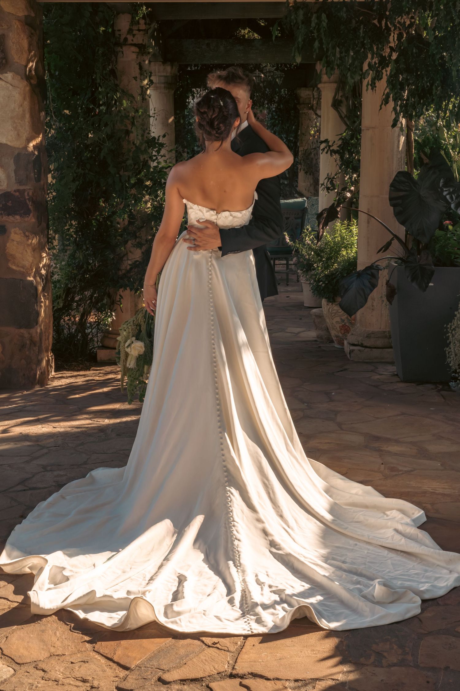 Bride in strapless gown with long train embracing groom in sunlit garden colonnade.