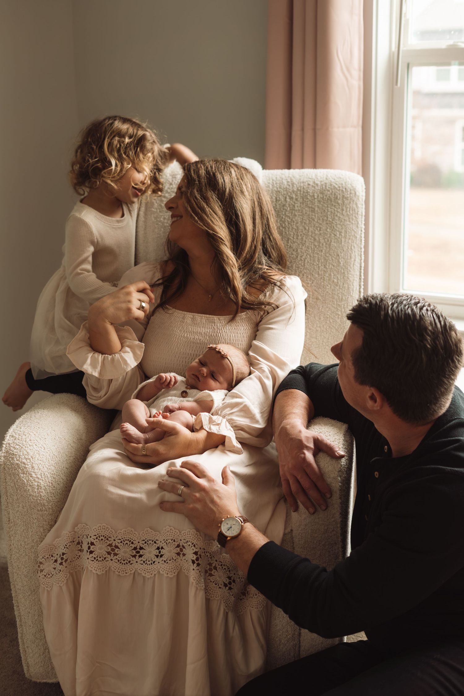 Family portrait with mother holding newborn in armchair, toddler beside her, father kneeling nearby by a bright window.