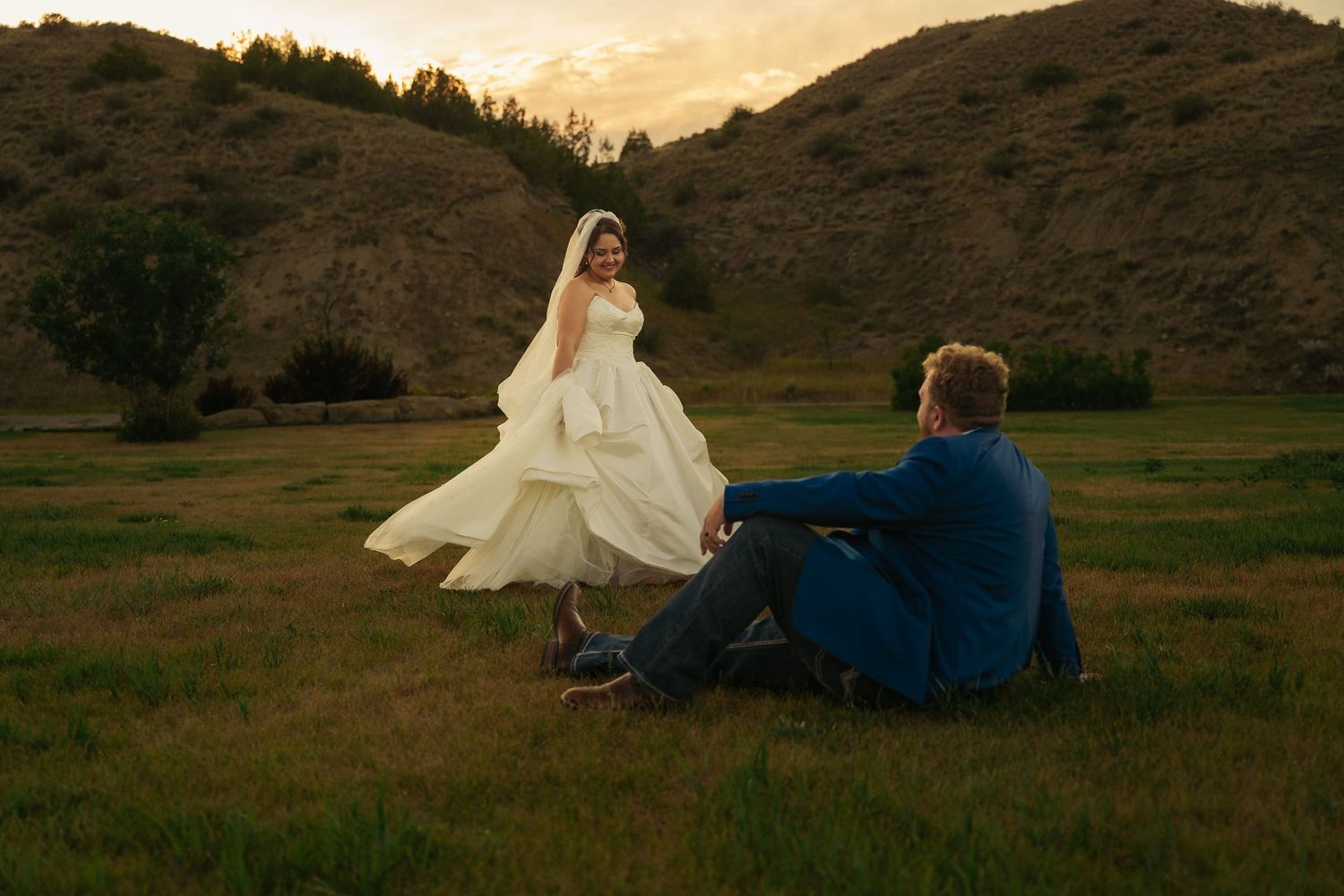 Bride in white gown twirling at sunset while groom sits on green grass with rolling hills backdrop.
