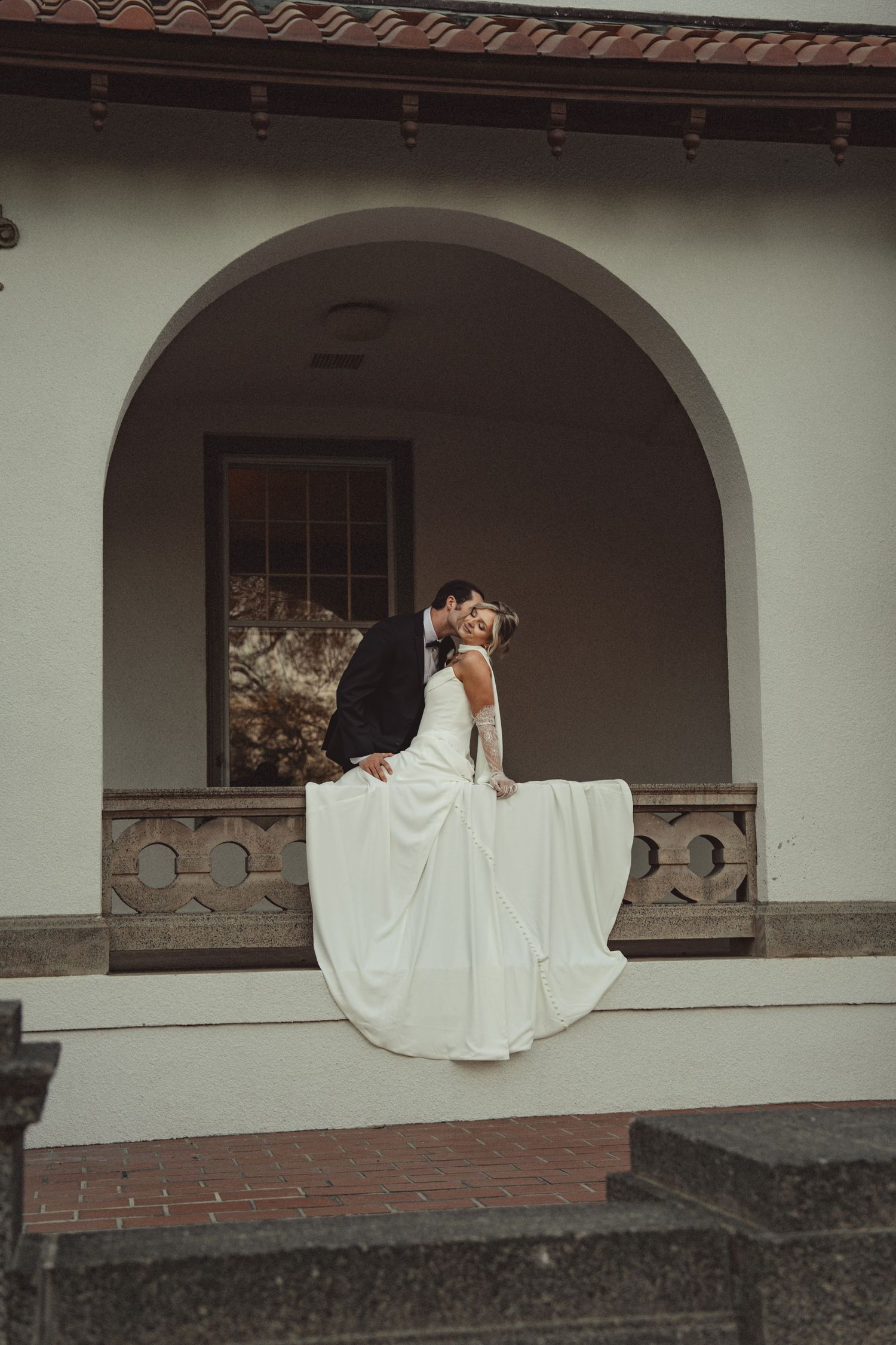 Bride and groom share a romantic kiss on a elegant white arched building porch, wedding dress flowing beautifully.